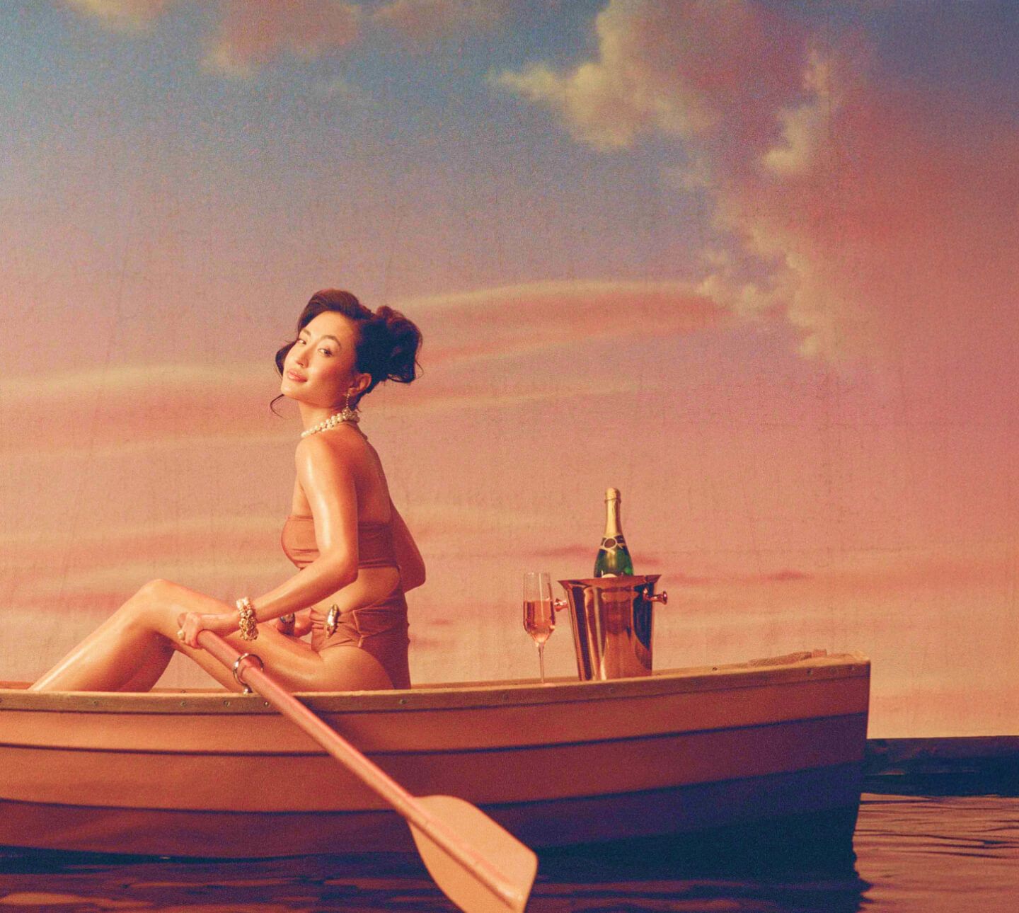 Smiling woman in a swimsuit in a rowboat with an oar, champagne, and a glass, under a pink sky.