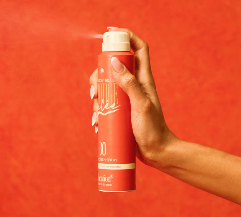 A hand with French-tipped nails sprays orange sunscreen mist from a can against an orange background.