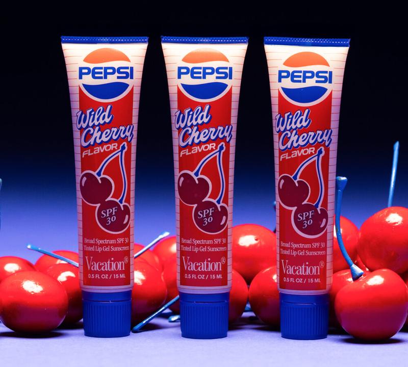 Three Vacation Pepsi Wild Cherry SPF 30 lip sunscreen tubes sit amidst shiny red cherries with blue stems.