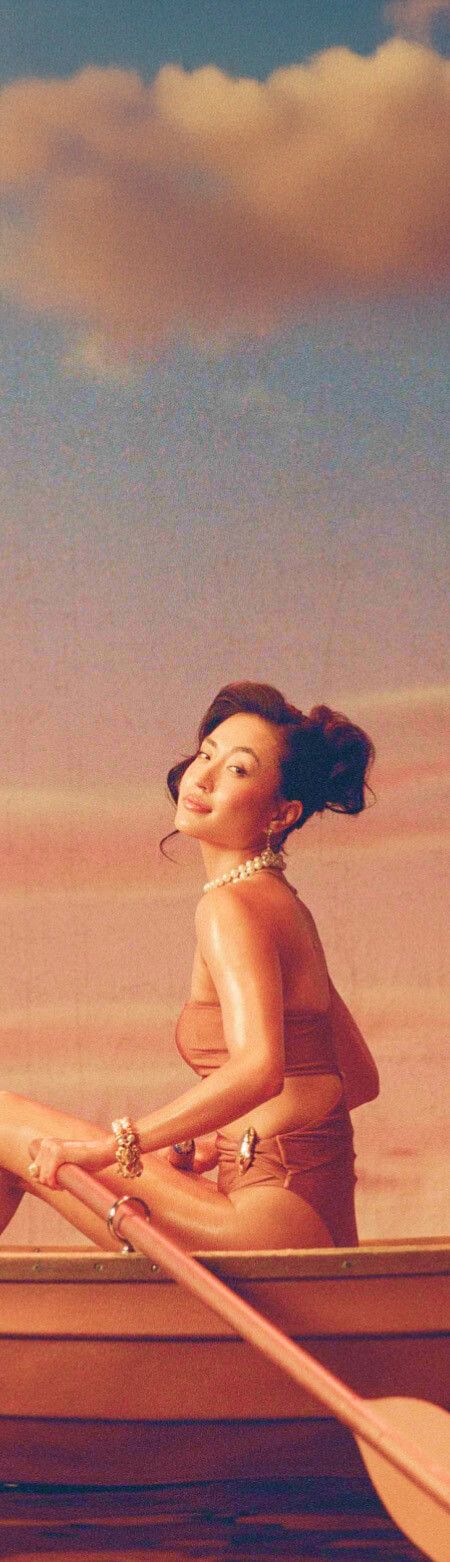Smiling woman in a swimsuit in a rowboat with an oar, champagne, and a glass, under a pink sky.