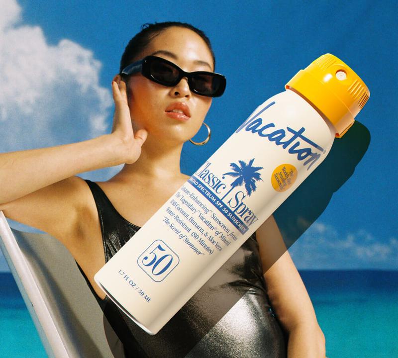 a woman wearing sunglasses is standing next to a bottle of vacation classic spray .
