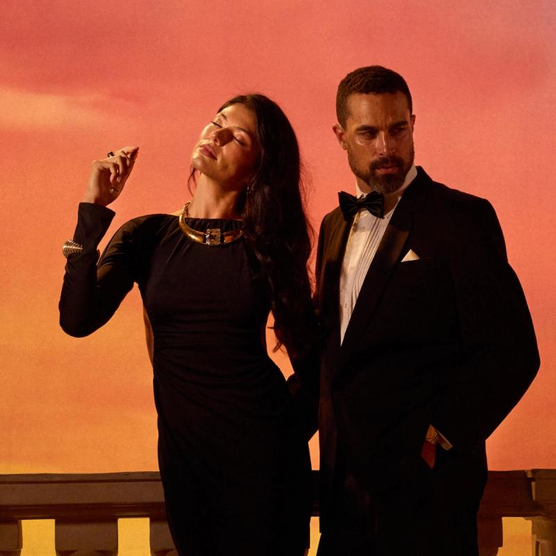 Woman in a black dress with a gold choker, head tilted back, next to a man in a tuxedo against an orange sunset.