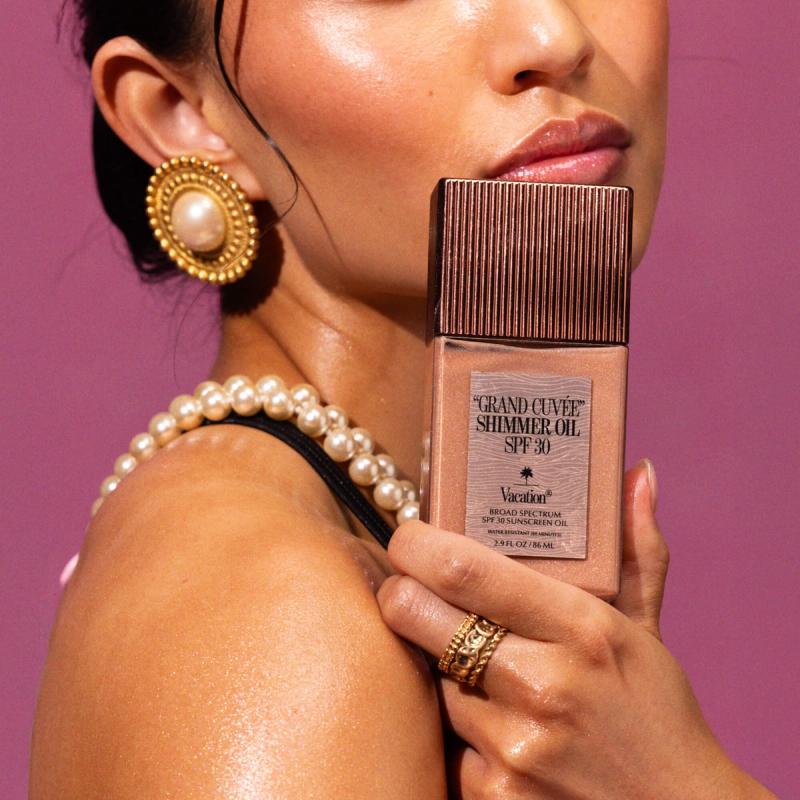 A person with shimmery skin, glossy lips, and pearl jewelry holds a bottle of Vacation Grand Cuvée Shimmer Oil SPF 30.