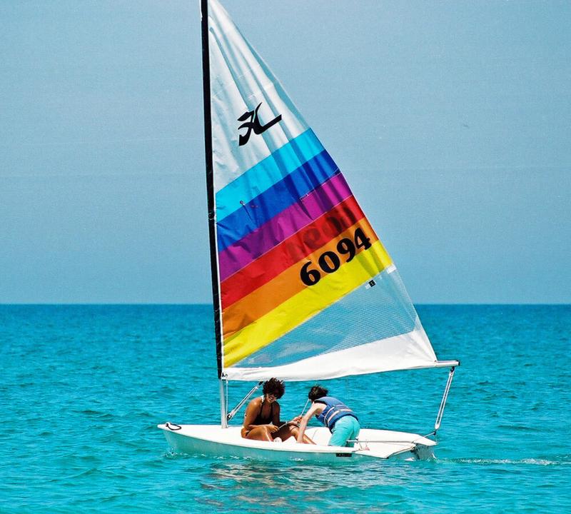Two people sailing a white Hobie Cat sailboat with a rainbow-striped sail on blue water.