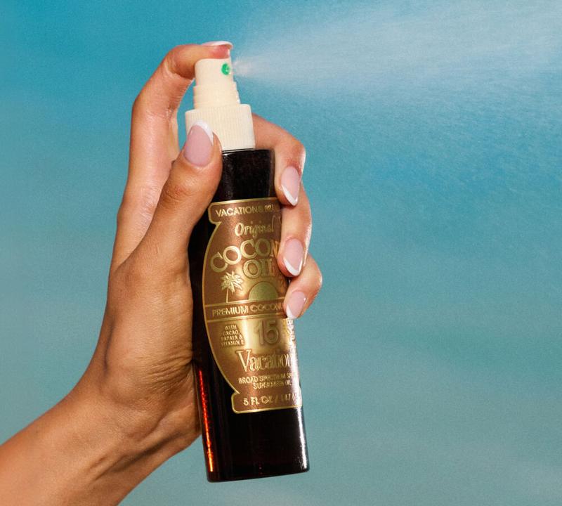 A hand sprays Vacation Inc. Original Coconut Oil SPF 15 from a dark bottle against a blue background.