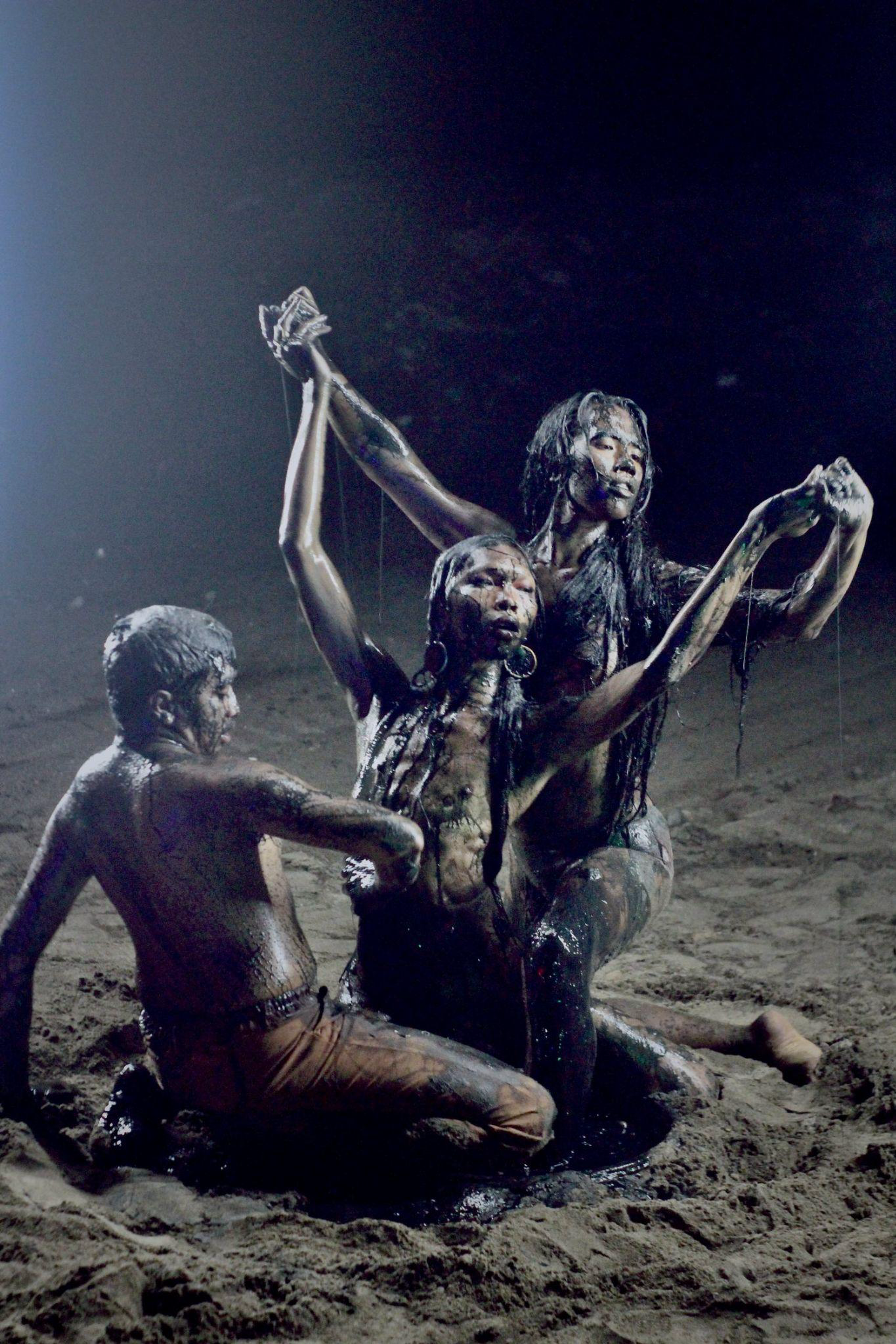 hree figures dance together on a beach at night time, seated and kneeling with arms interconnected and raised towards the sky. They are covered in dark earth.