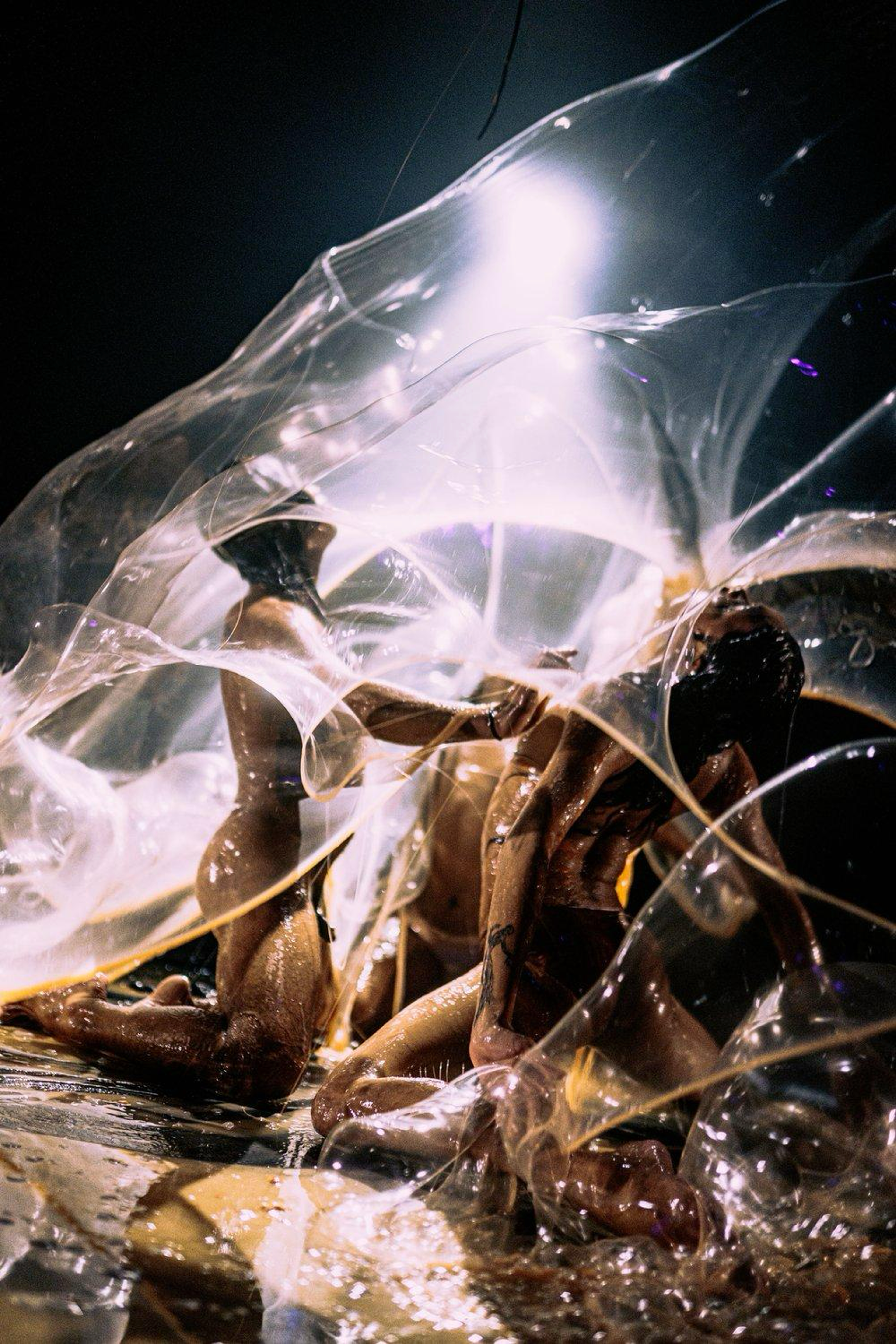 A group of figures move together under layers of transparent, latex-like material. Light, liquid, and textures blur together into abstraction under a spotlight in an otherwise dark room