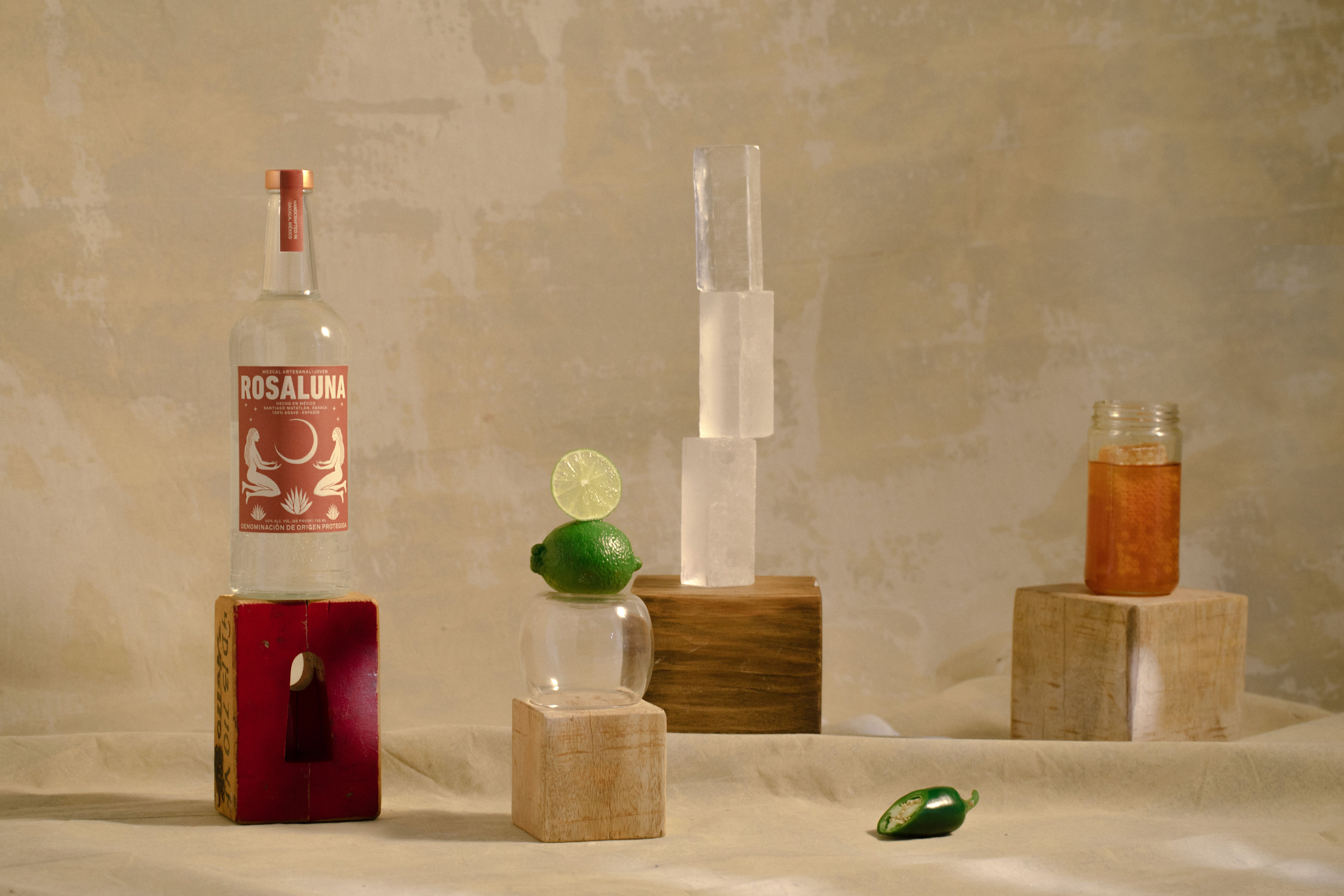 Still life composition of a Rosaluna Mezcal bottle and various objects, fruits, and wooden blocks.
