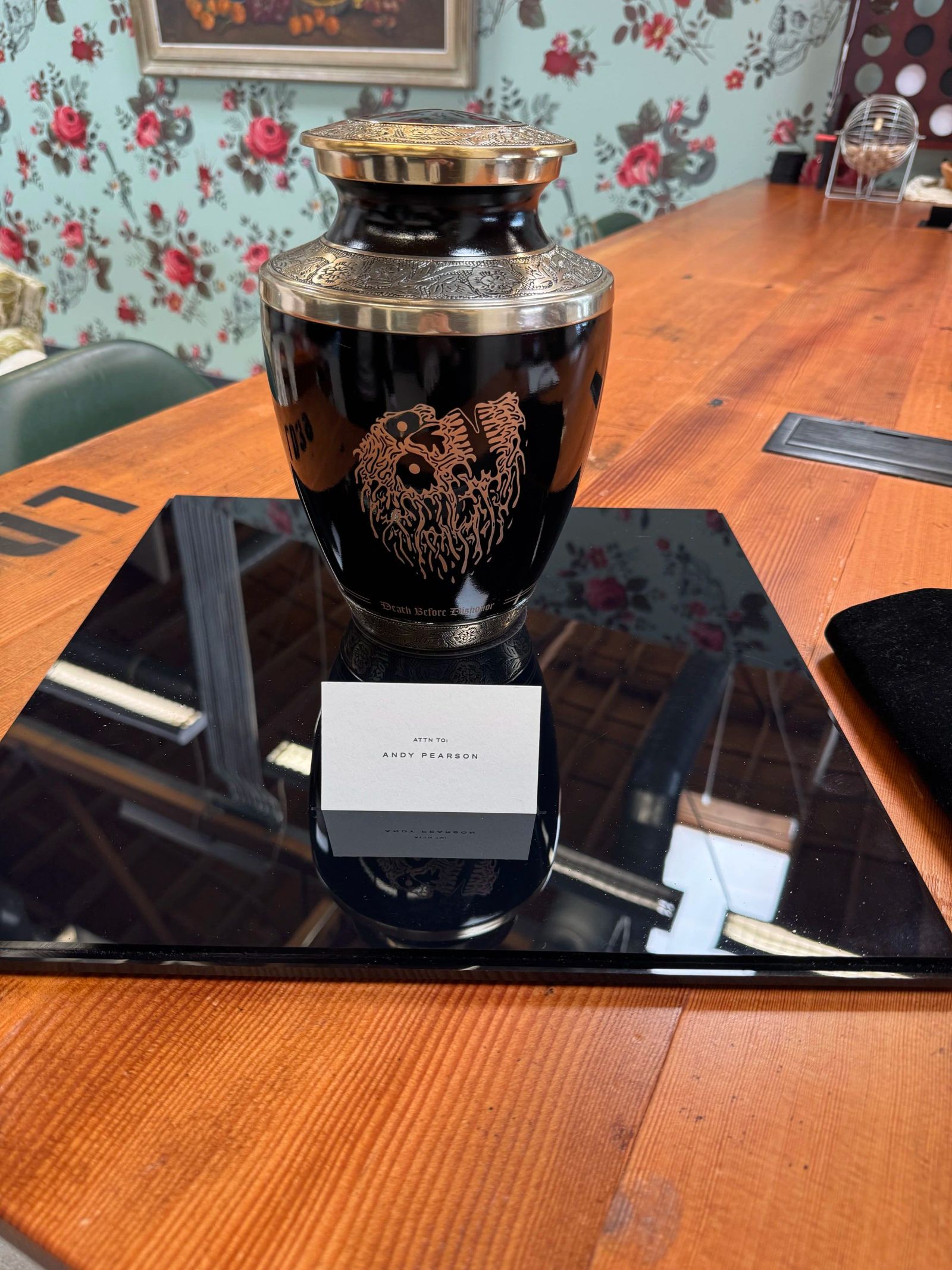 A black and silver urn engraved with the Liquid Death skull, with a place card addressed to Andy Pearson, on a wooden table
