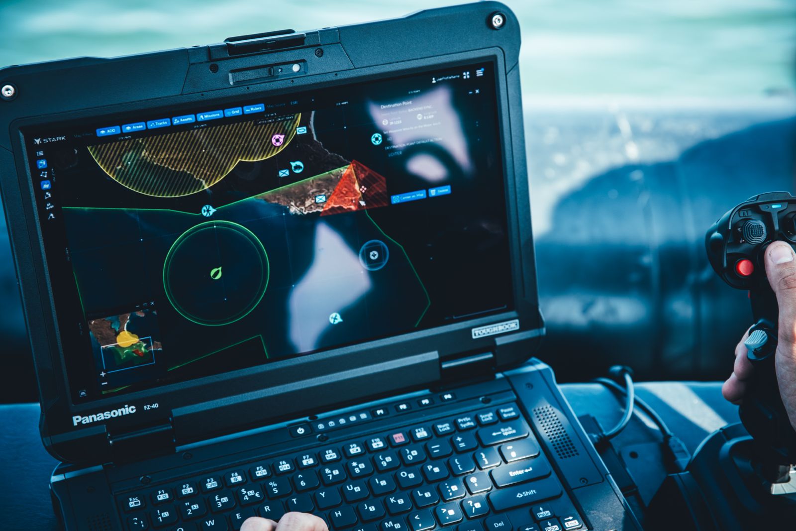 Hand operating a joystick next to a rugged laptop displaying a drone control interface with maps and waypoints.