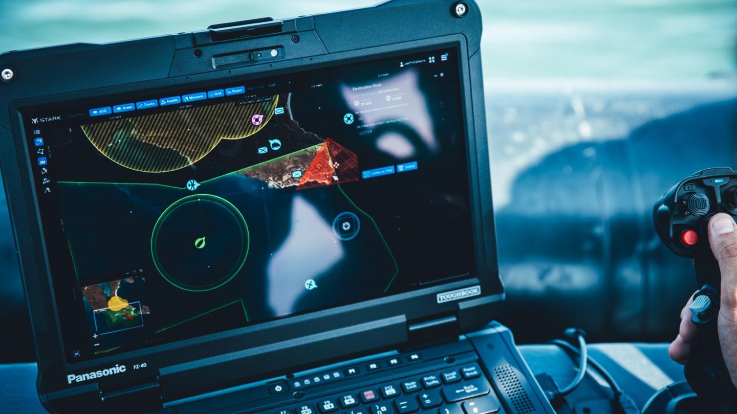 Hand operating a joystick next to a rugged laptop displaying a drone control interface with maps and waypoints.