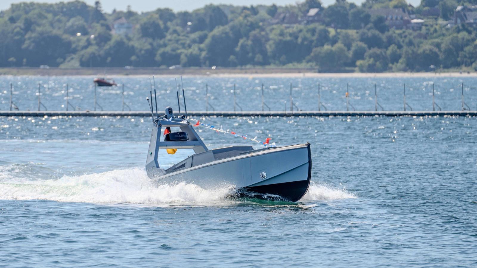 A gray autonomous boat moves quickly across the water, leaving a white wake, with a distant treed shoreline and a floating barrier in the background.