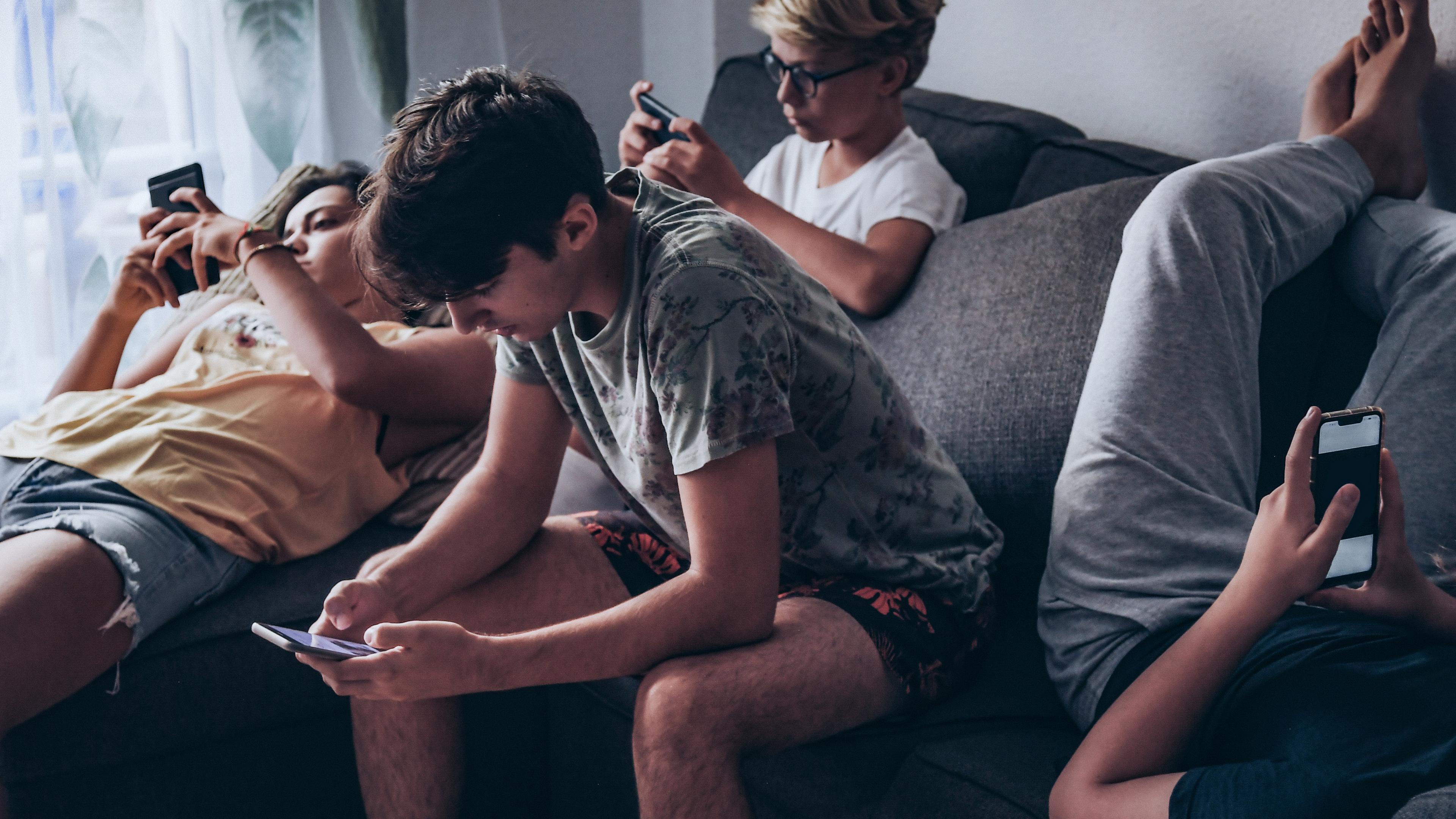 Fire unge drenge slænger sig i en sofa, mens de kigger på hver sin telefon.
