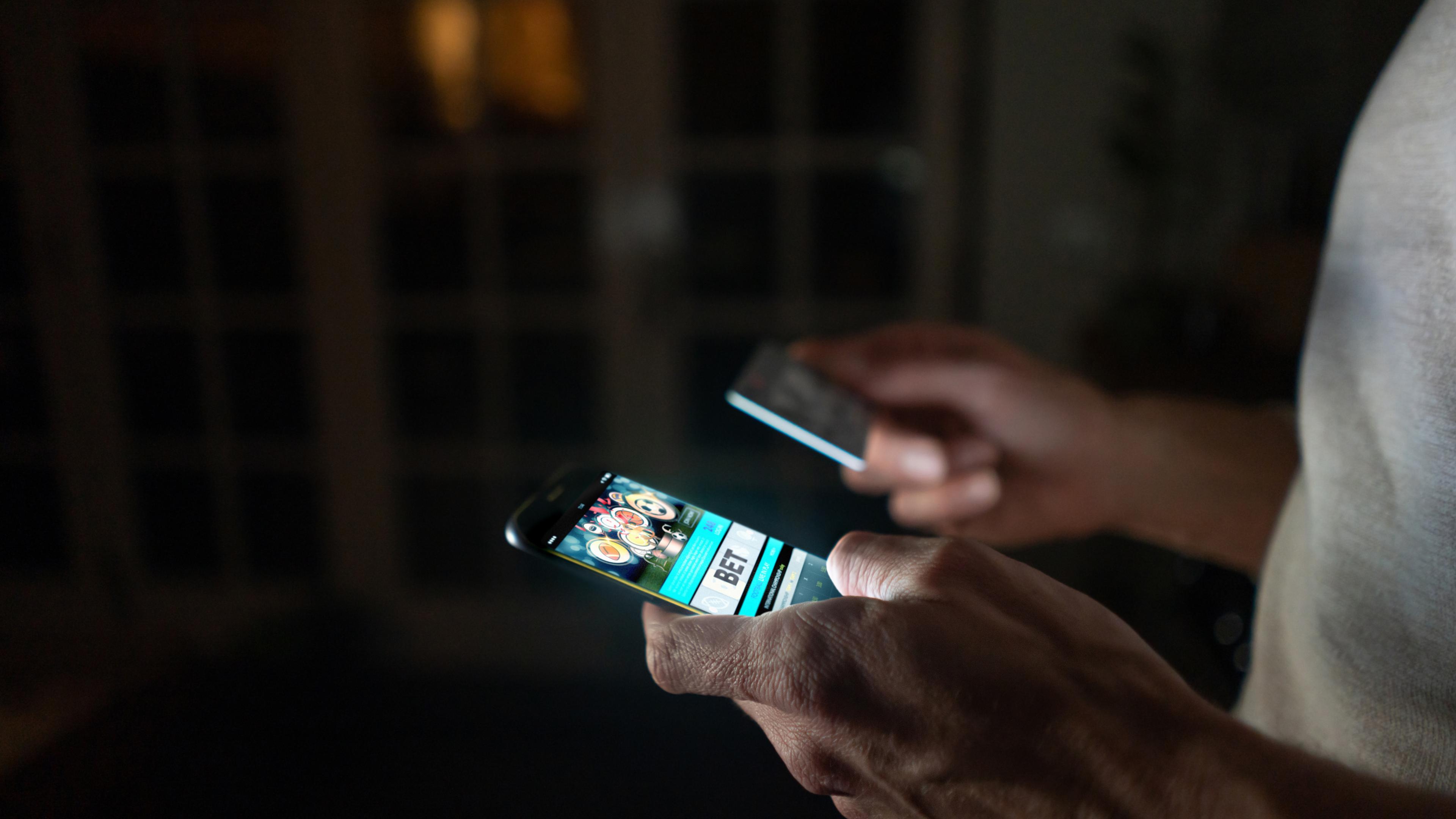 Close-up af en mand, der står i et mørkt rum med sin mobiltelefon i den ene hånd og et betalingskort i den anden. På skærmen kan man se, at han er ved at placere et væddemål på en spilside. 