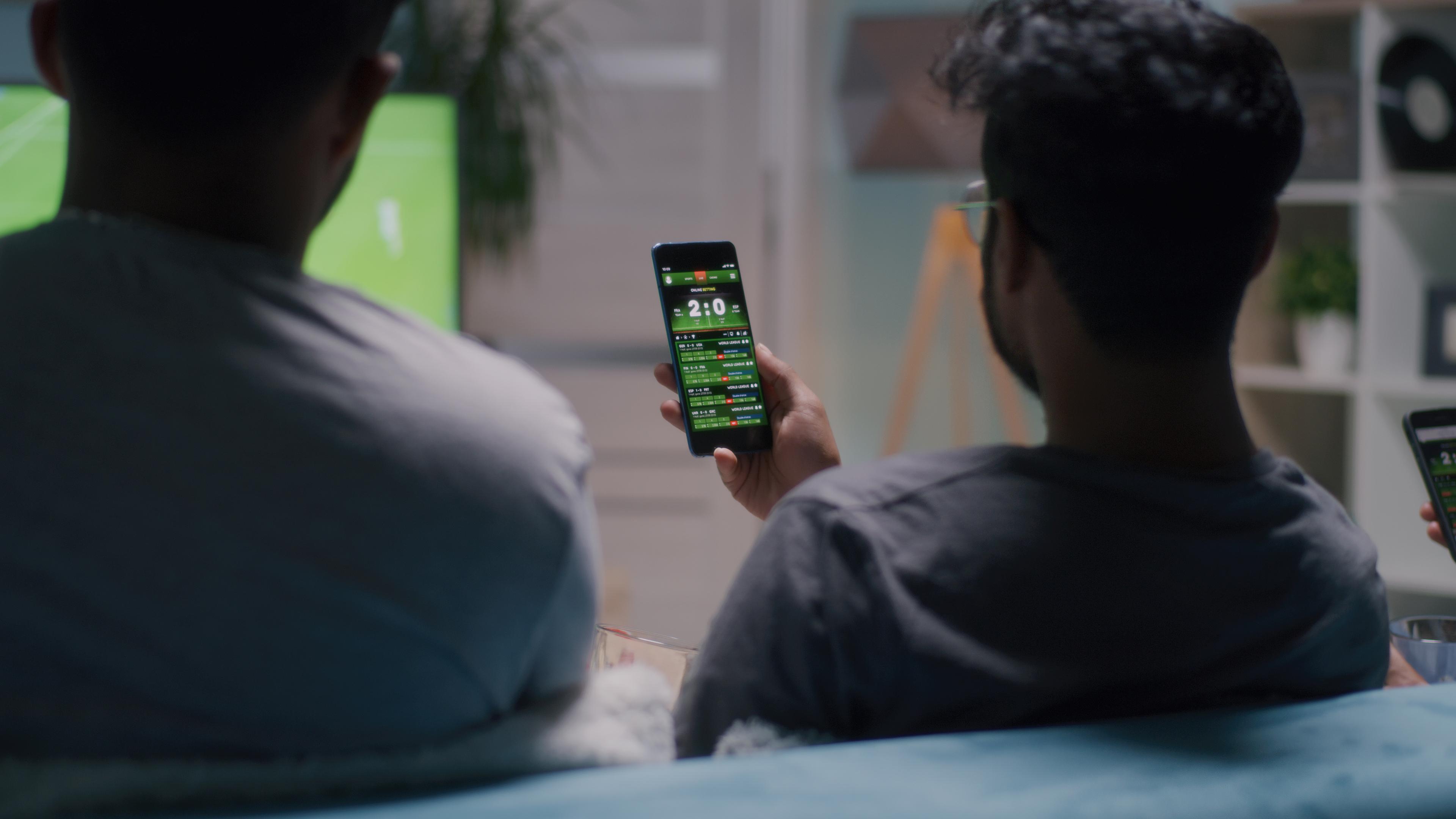 To personer oddser på mobilen, mens de ser en fodboldkamp på fjernsynet