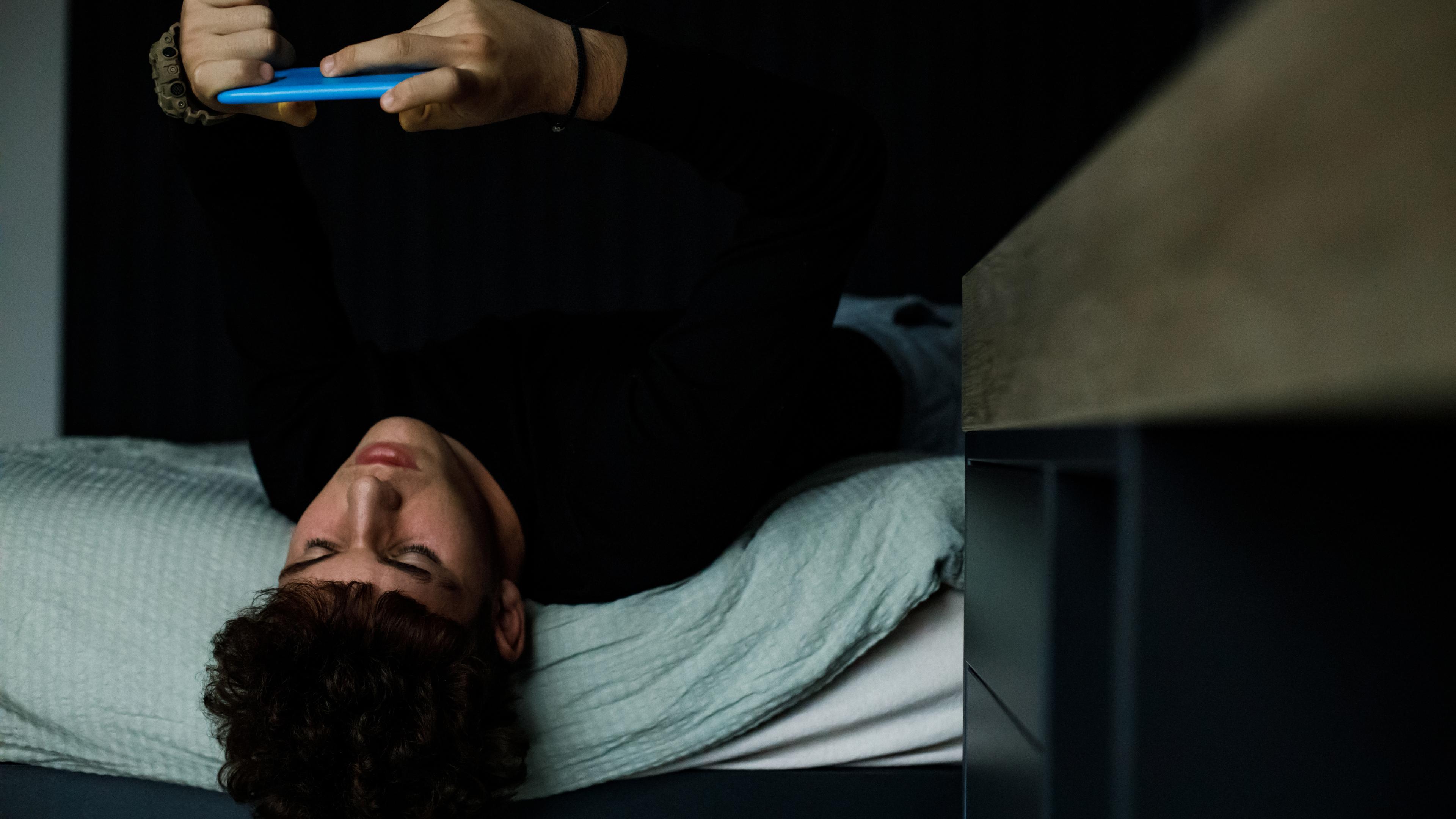 Dreng ligger på sin seng med hovedet nedad og kigger på sin telefon.