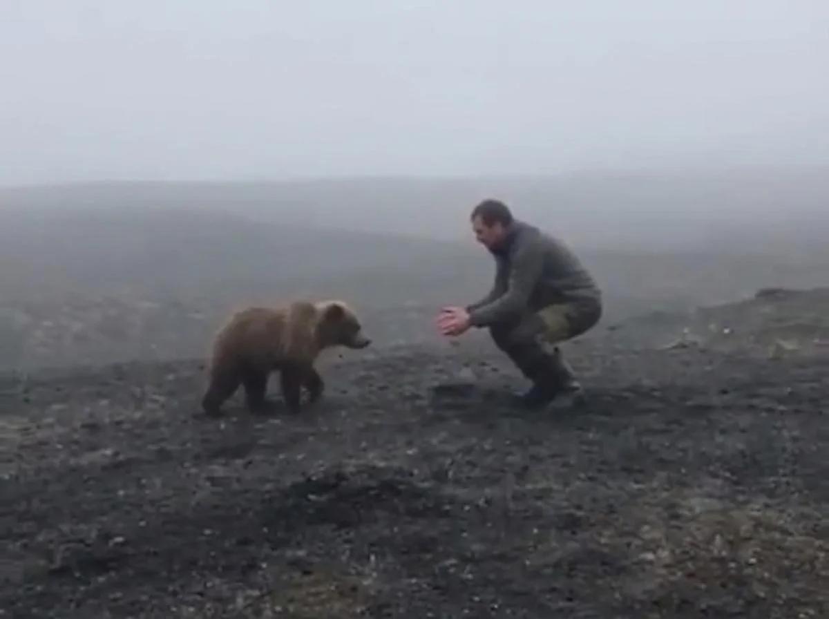 A blurry, foggy image of a man bending down as a small baby bear clings to his leg.