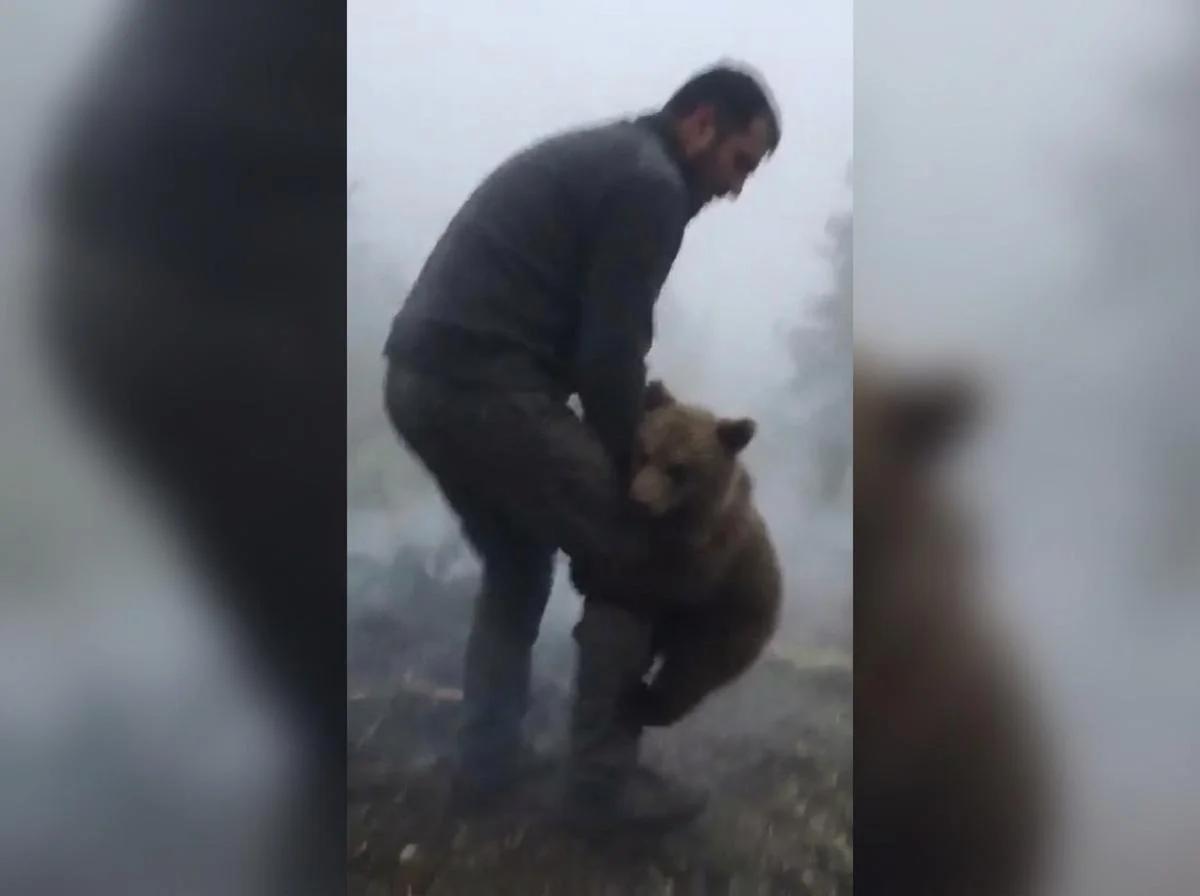 A man in a foggy setting crouches down and holds out his hands to a small baby bear.