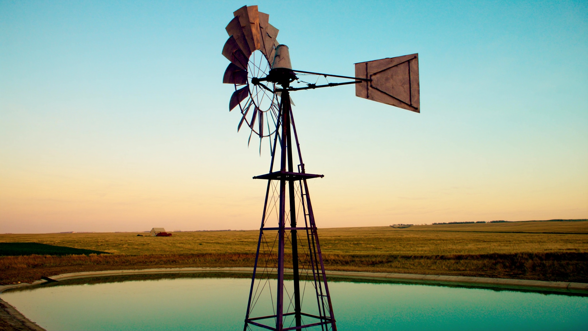 A classic multi-blade farm windmill aerator beside a calm prairie dugout pond at golden hour