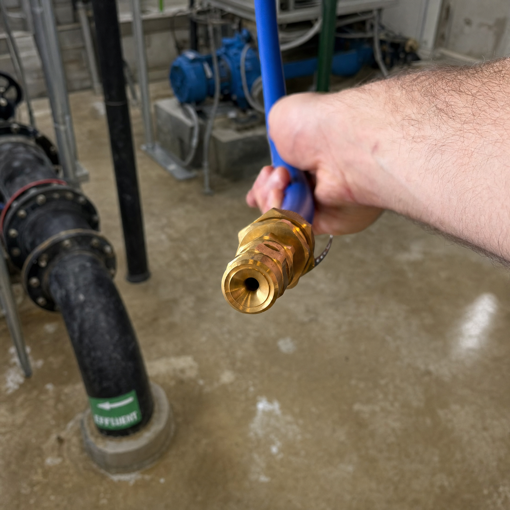 A brass dispensing nozzle on a blue hose, held above a lift-station effluent pipe