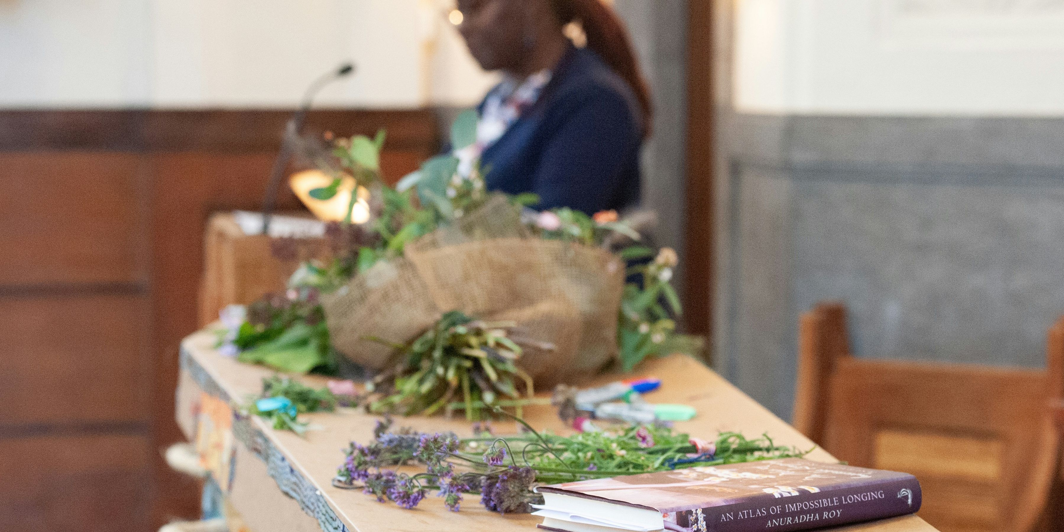 A woman stands beside a cardboard coffin in a crematorium chapel. On top of the coffin is a bouquet of flowers and a book.