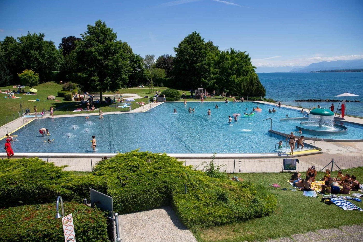 Vista das piscinas exteriores de Colovray em Nyon, com o Lago Léman e os Alpes ao fundo, num dia de sol com muitos banhistas.