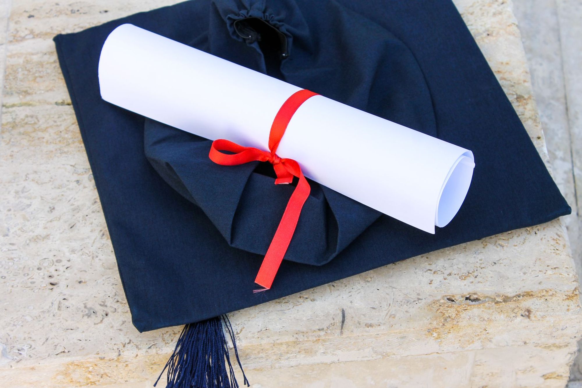 Navy graduation cap with a rolled certificate tied with a red ribbon, resting on a pale stone ledge