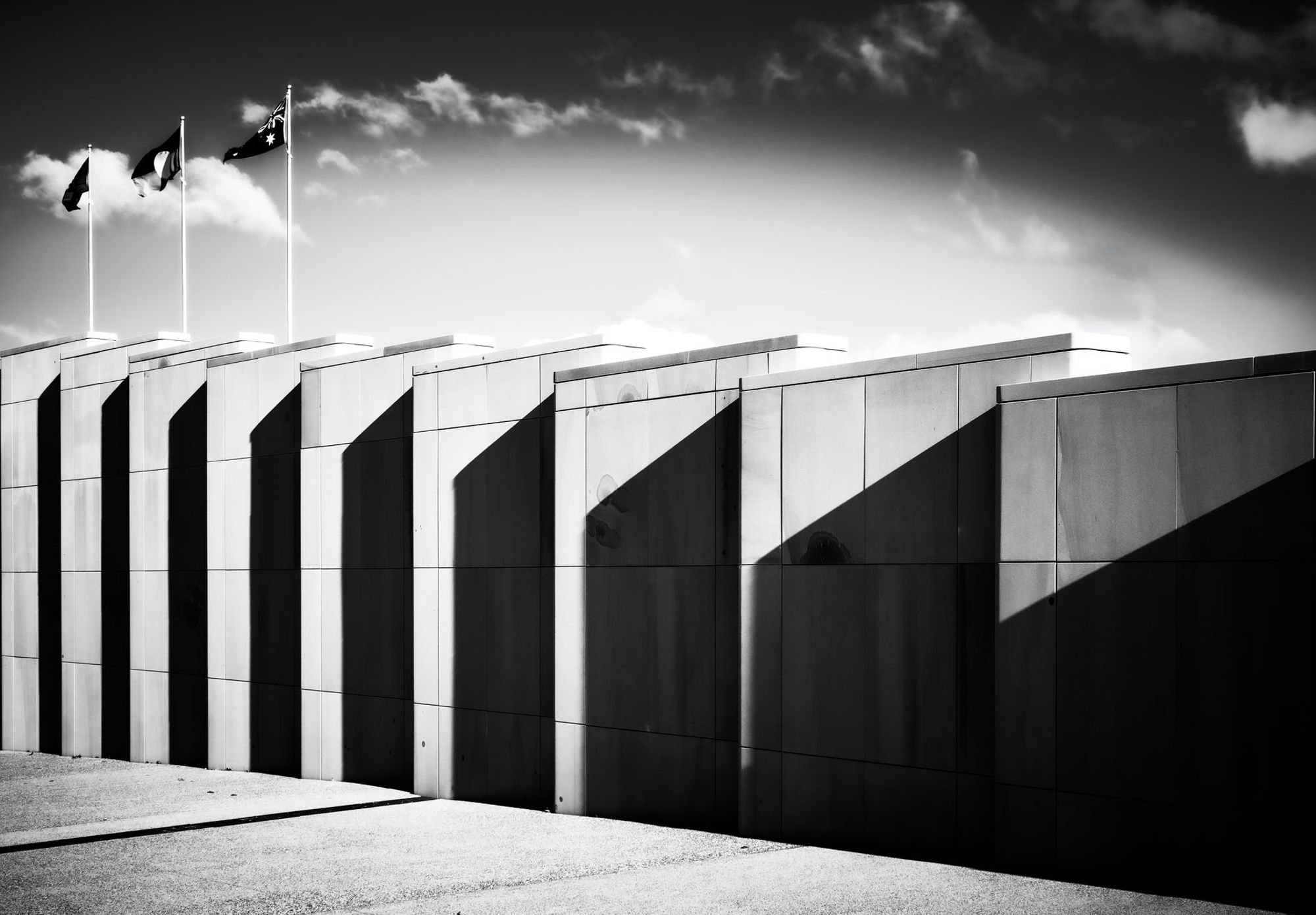 Black and white photo of sandstone walls in Canberra casting long shadows, with Australian flags flying above