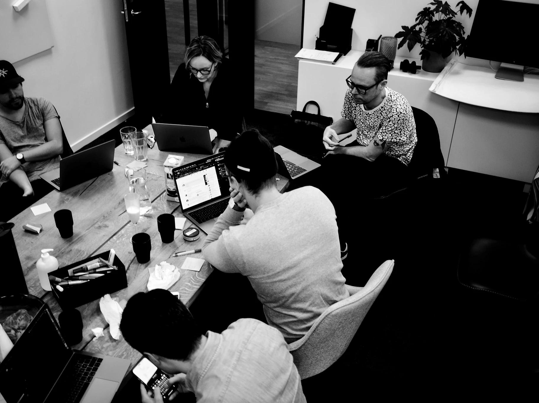 Fotografi taget med fågelperspektiv på fem personer som sitter vi arbetsbord, arbetar vi datorer. På bordet står kaffekoppar, servetter, pennor.