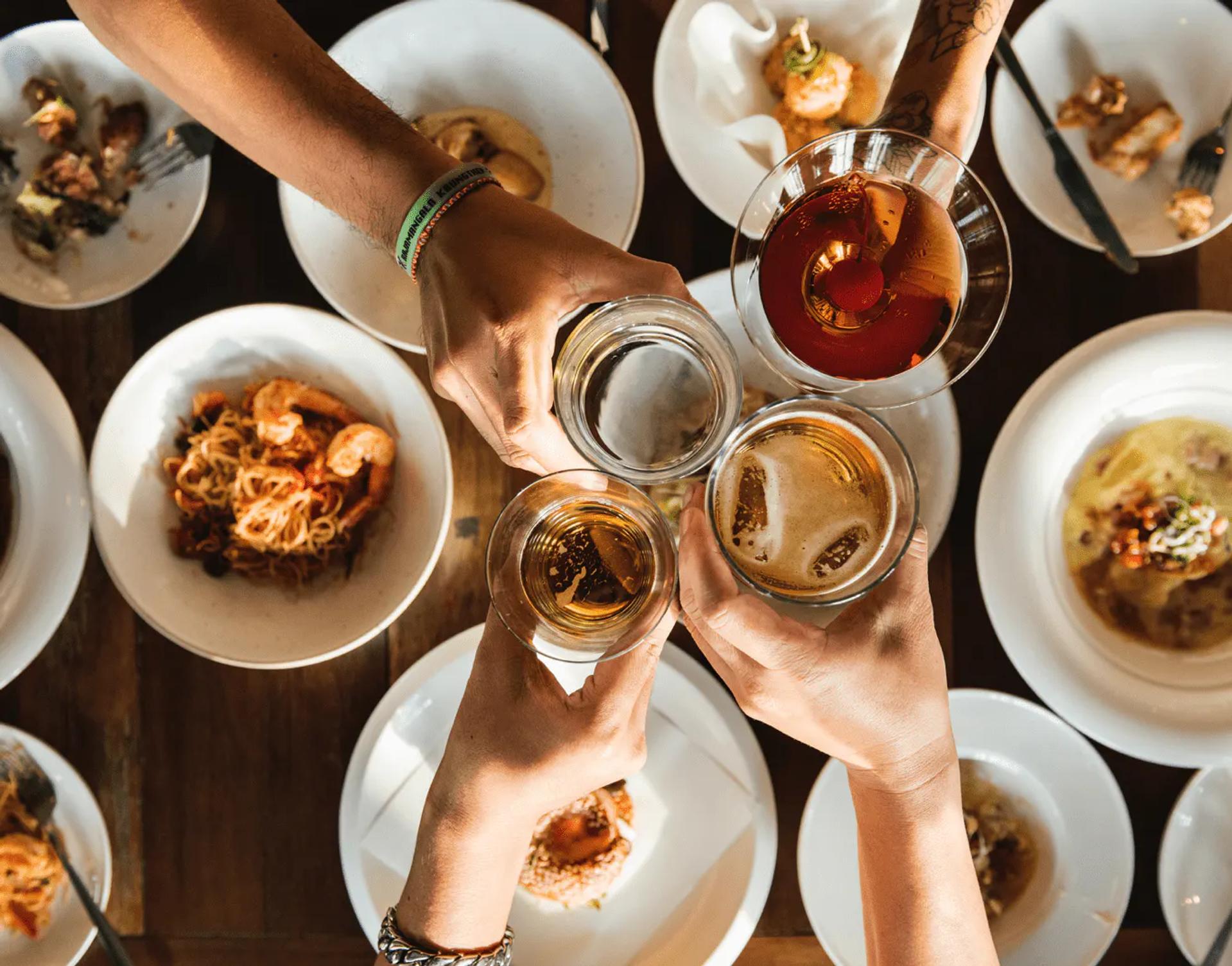 A table with multiple plates of food and hands cheersing with glasses of wine