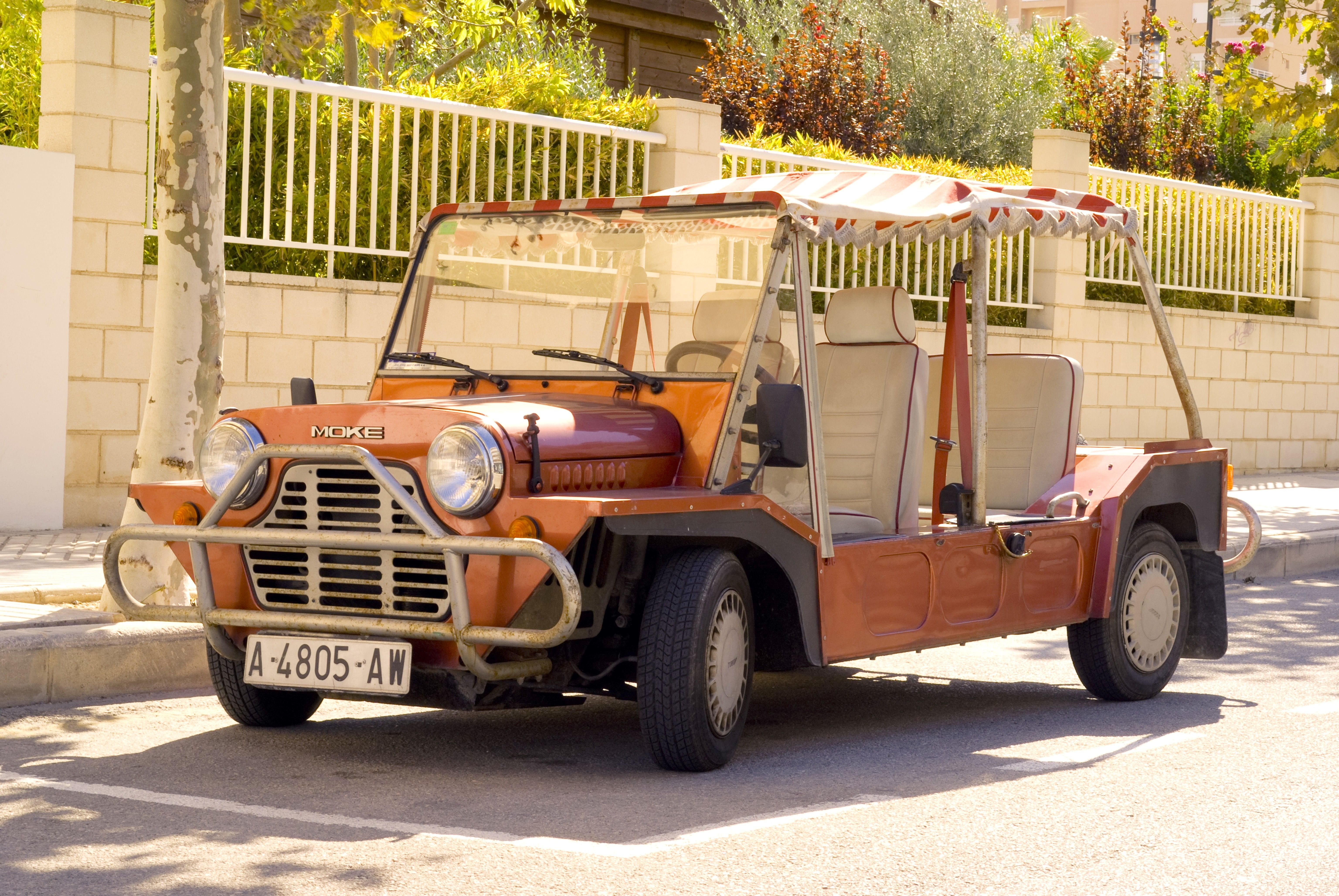 A vintage open-top Moke vehicle parked on a sunny street in a quiet neighborhood