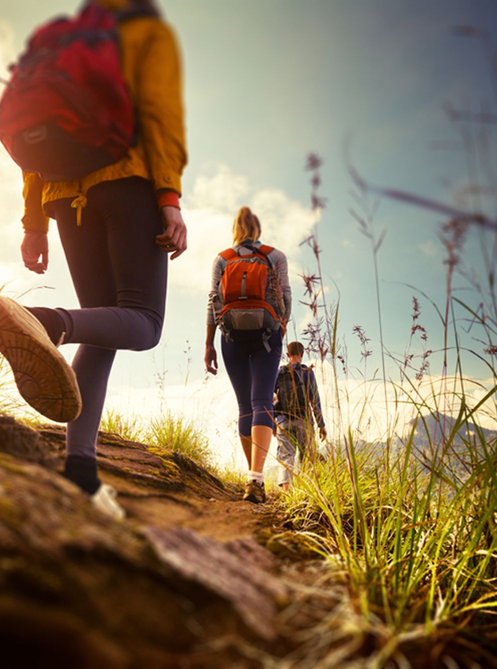 Group of hikers walking along a narrow mountain trail during sunset, with grassy surroundings and backpacks on