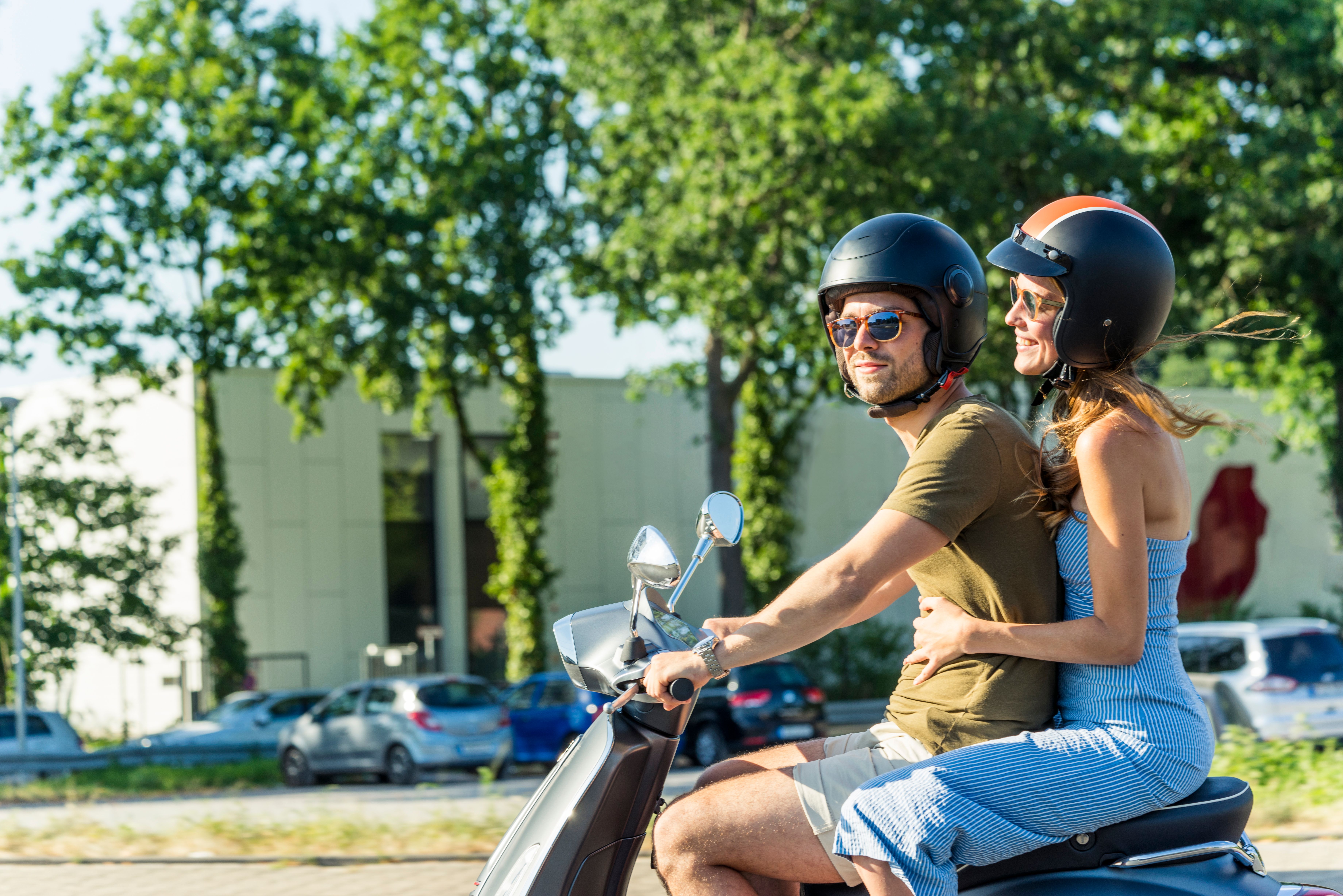 Smiling couple riding a scooter through a sunny city street, both wearing helmets and sunglasses