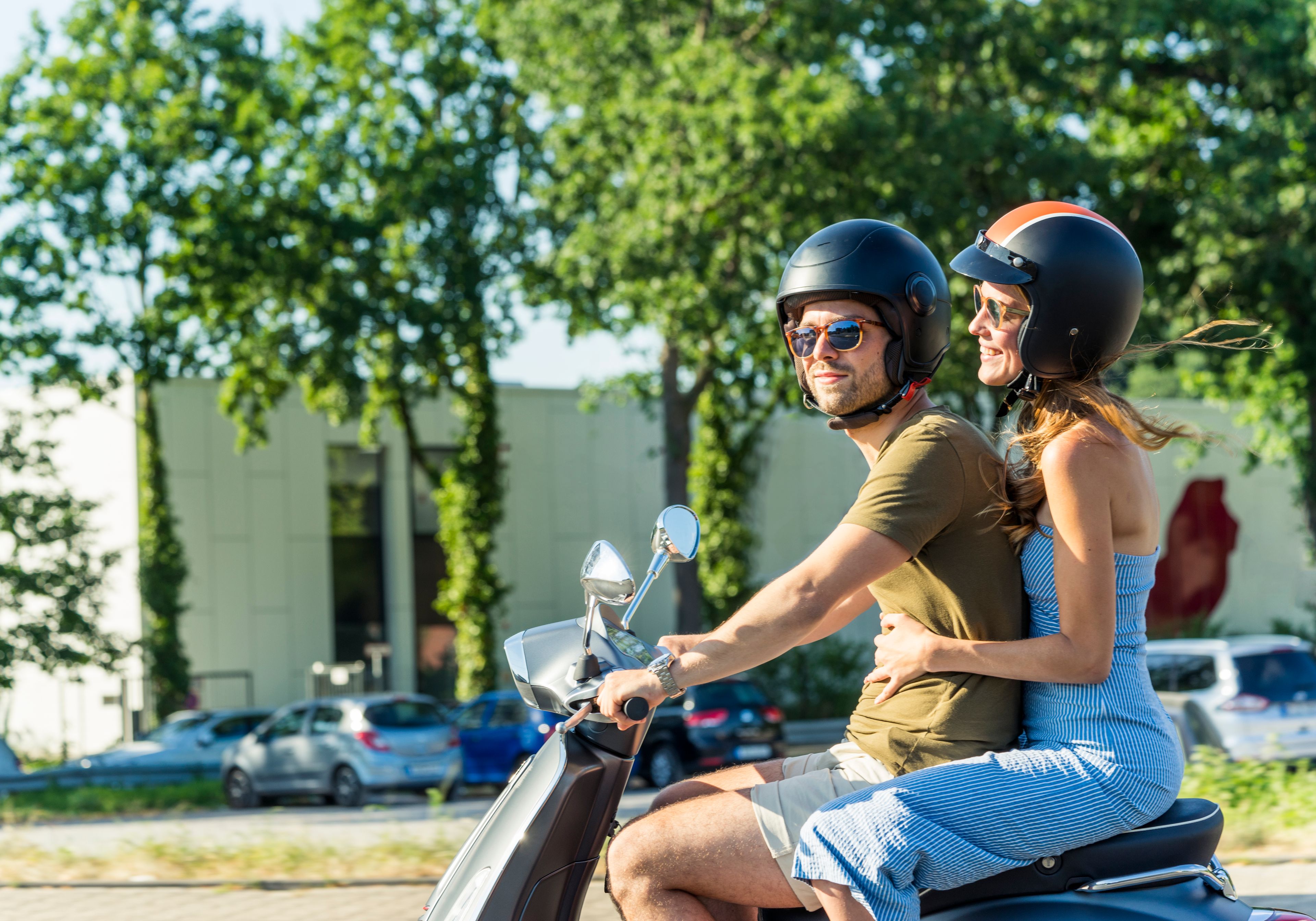 Smiling couple riding a scooter through a sunny city street, both wearing helmets and sunglasses