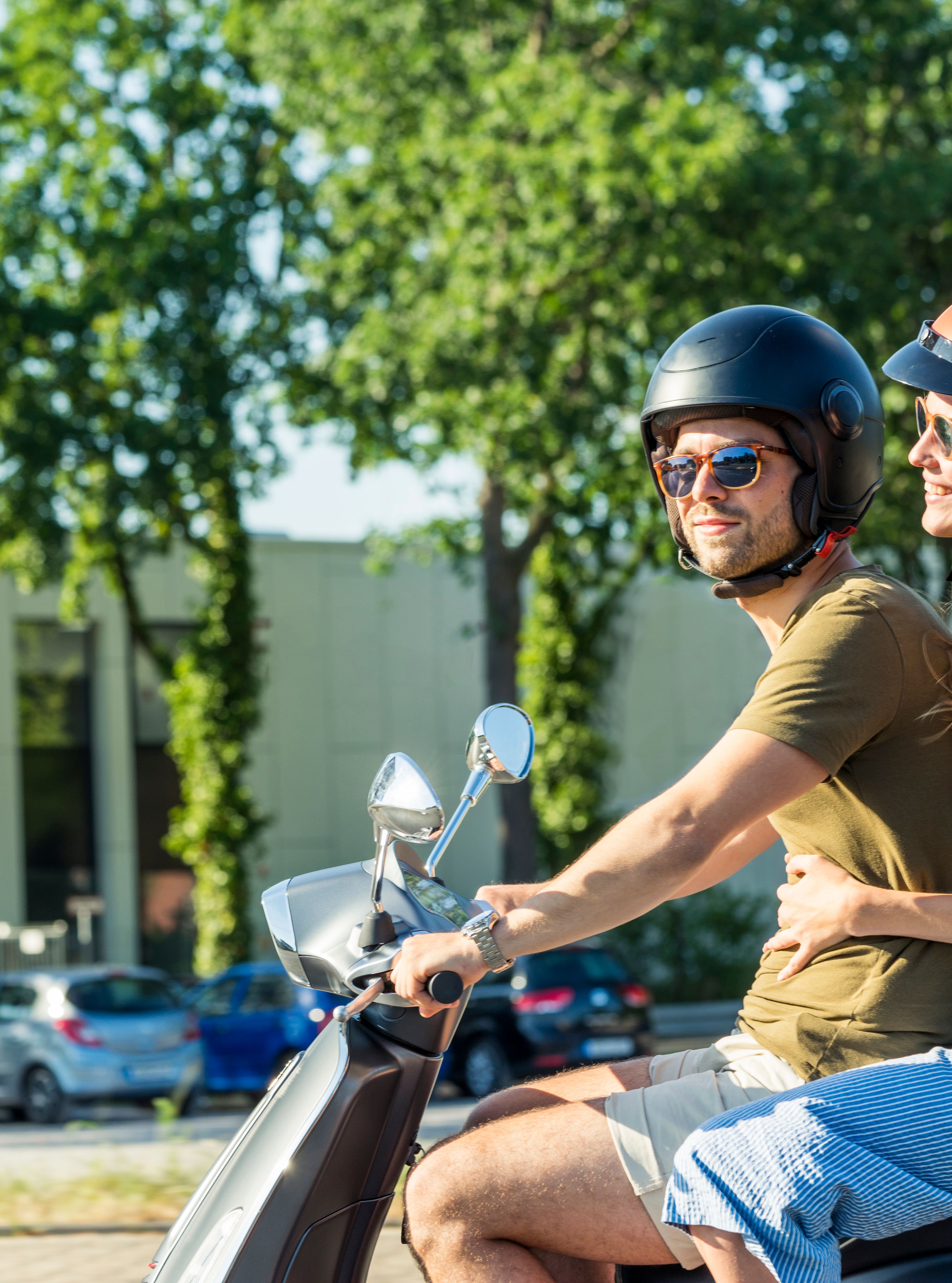 Smiling couple riding a scooter through a sunny city street, both wearing helmets and sunglasses