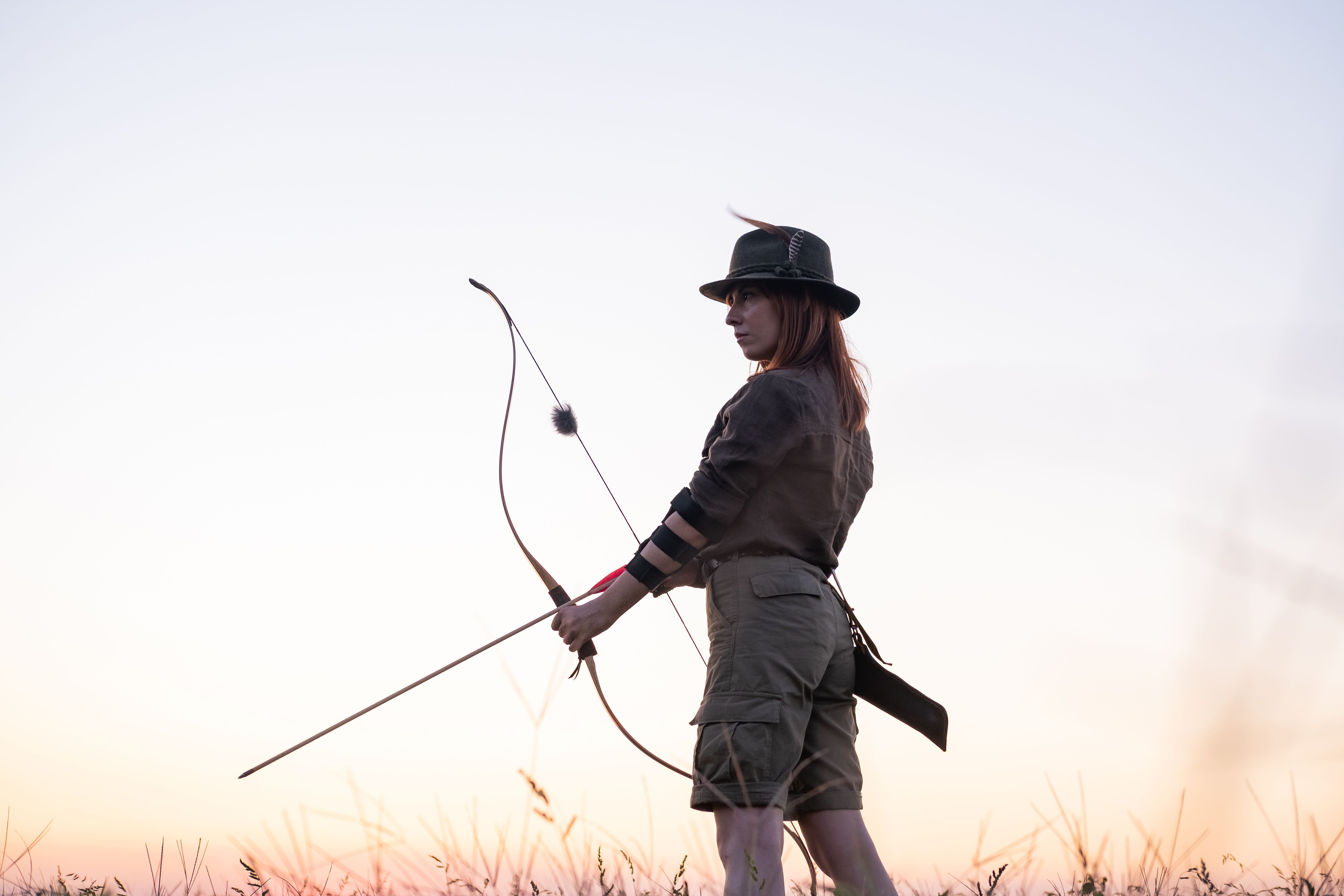 Woman standing in a field at sunset, wearing a hat and holding a bow and arrow, prepared for archery