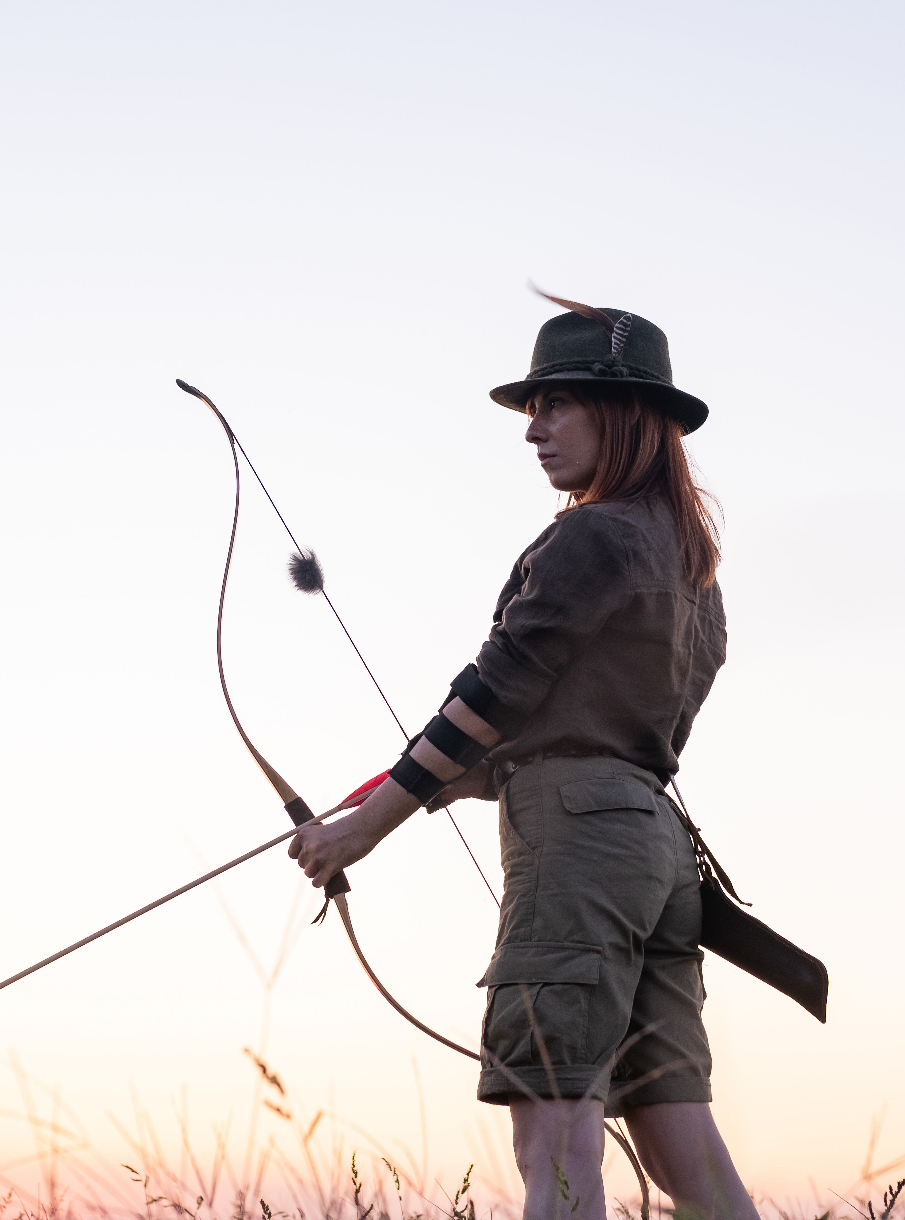 Woman standing in a field at sunset, wearing a hat and holding a bow and arrow, prepared for archery