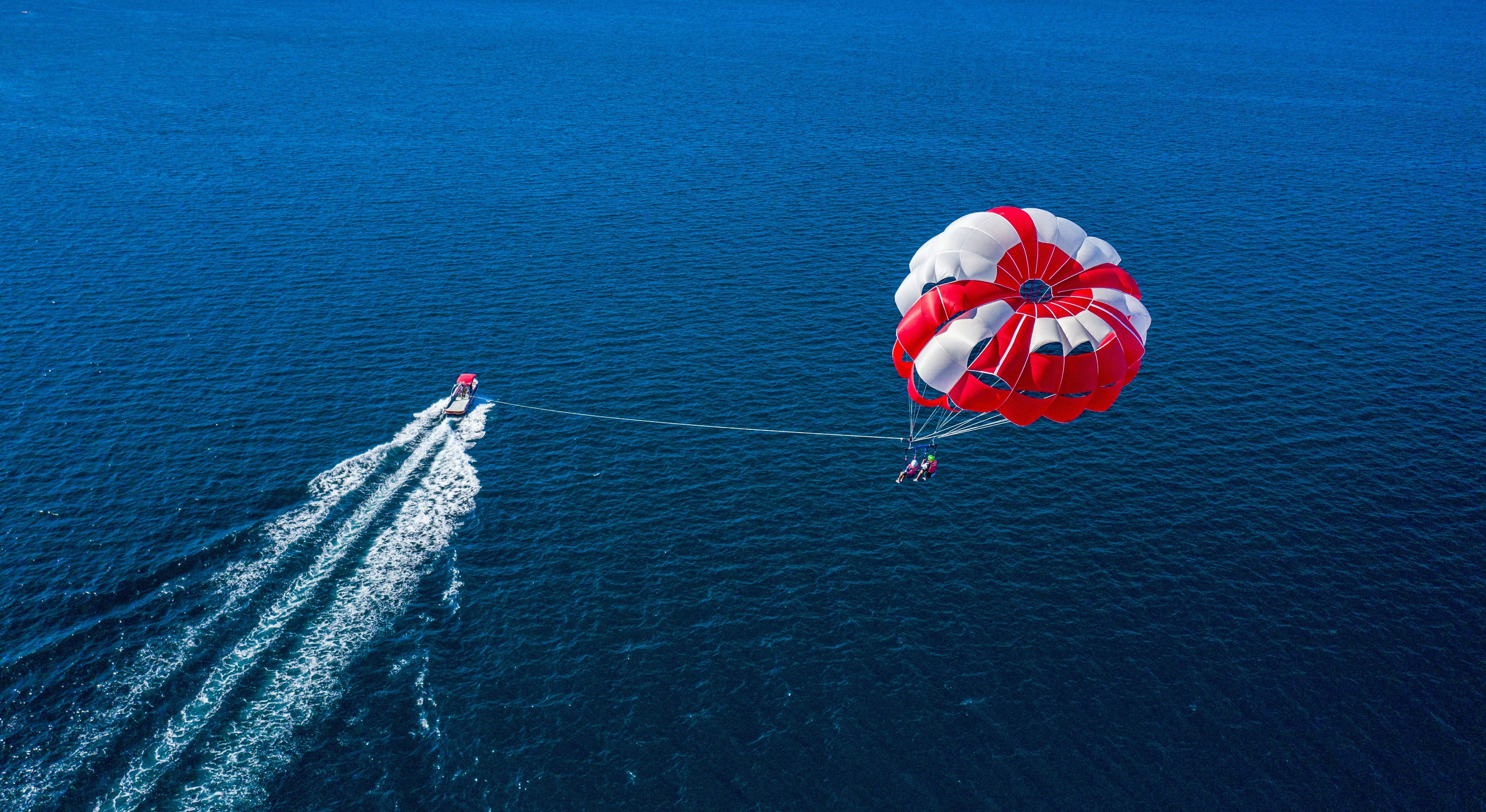 Aerial view of a boat pulling a person parasailing high above the open ocean under a large red and white parachute