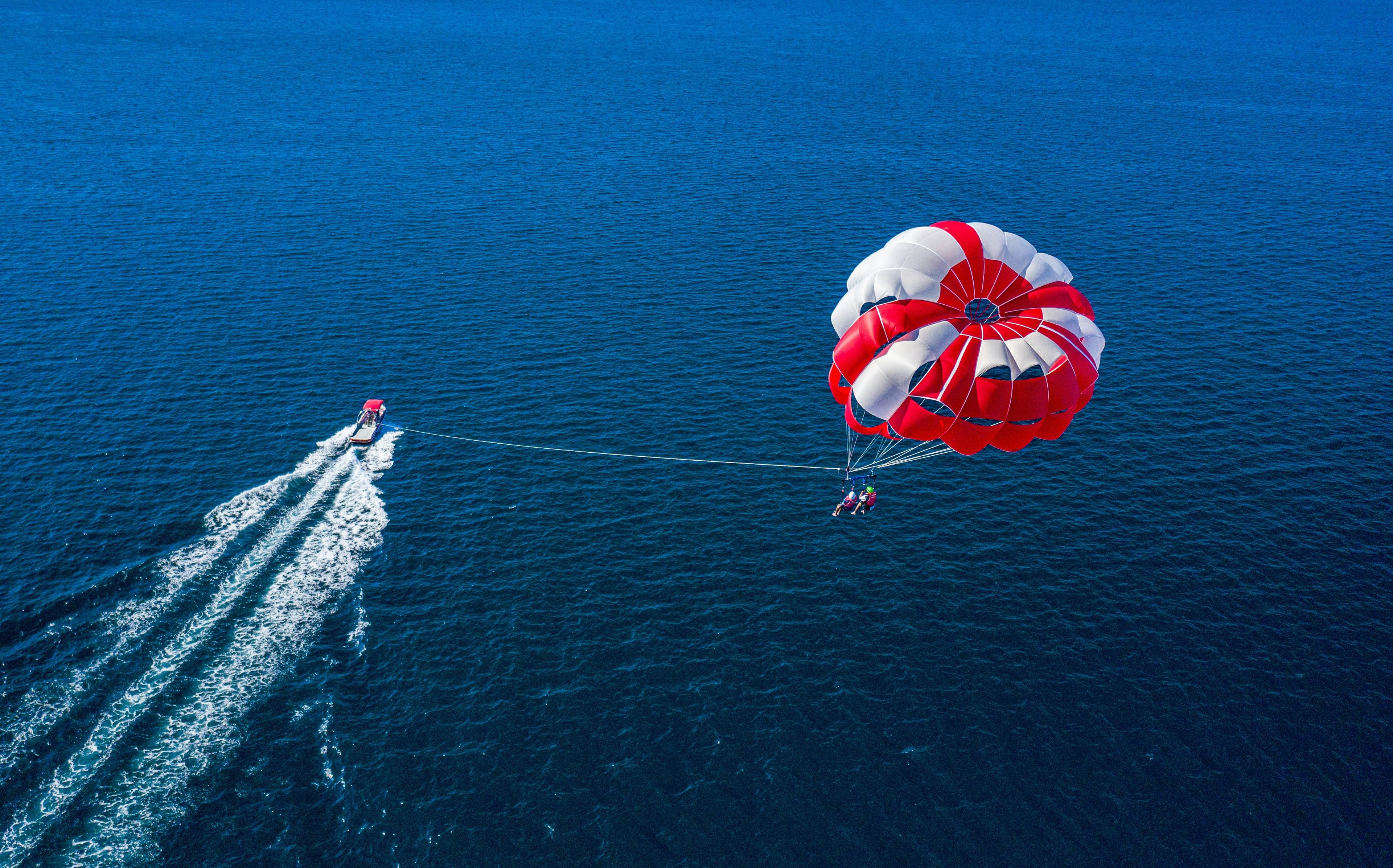 Aerial view of a boat pulling a person parasailing high above the open ocean under a large red and white parachute
