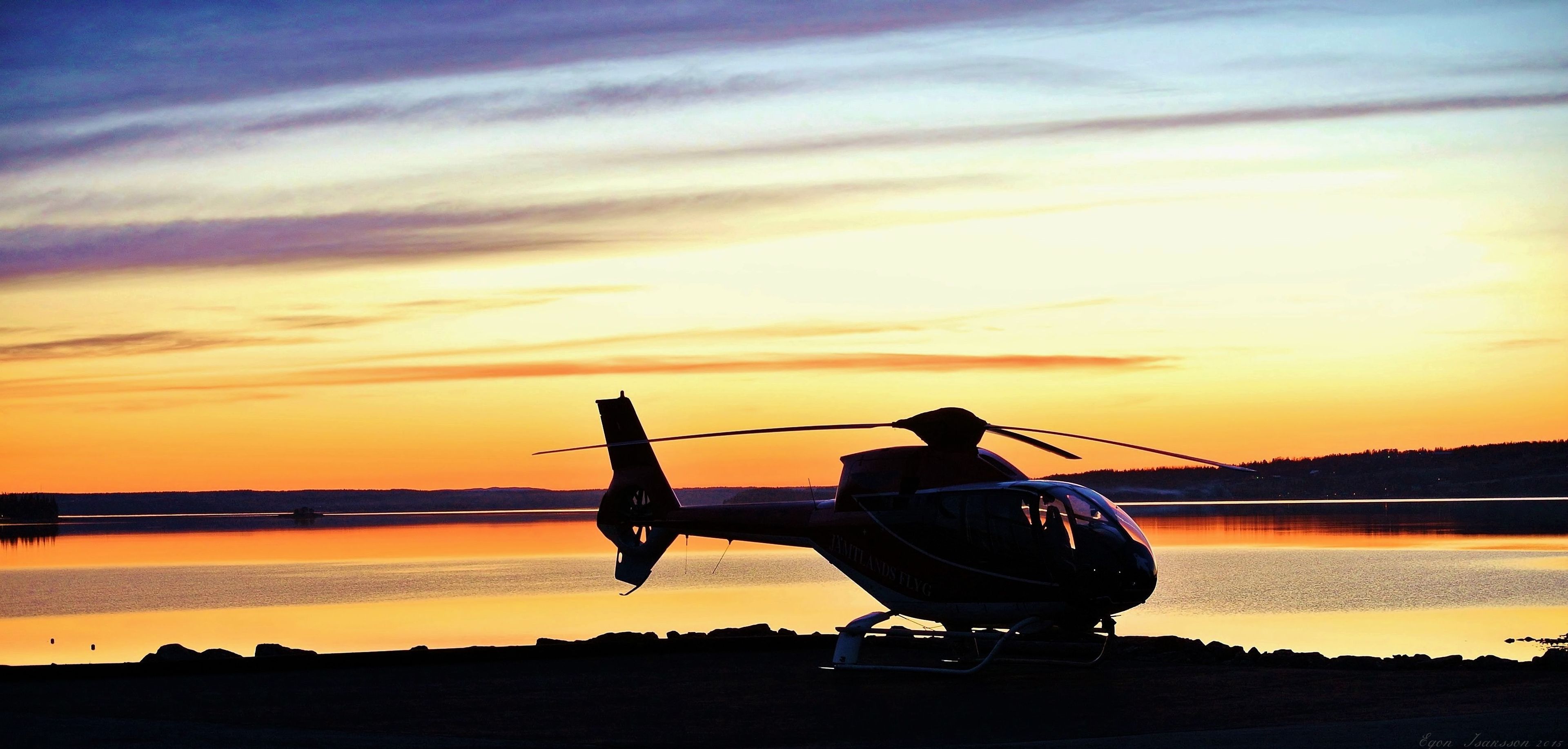 Helicopter parked near a lake during a colorful sunset, silhouetted against the sky reflecting on the water