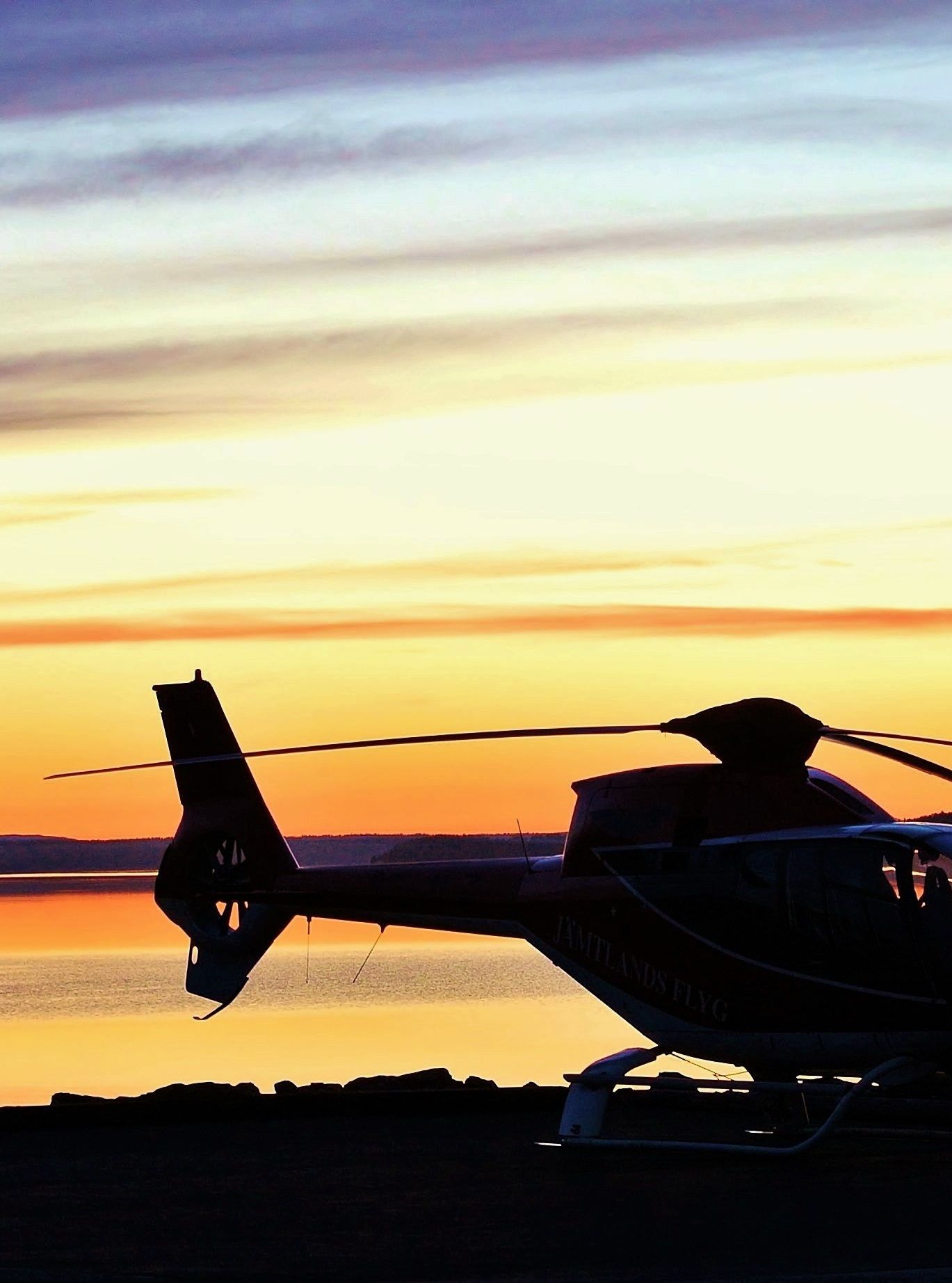 Helicopter parked near a lake during a colorful sunset, silhouetted against the sky reflecting on the water
