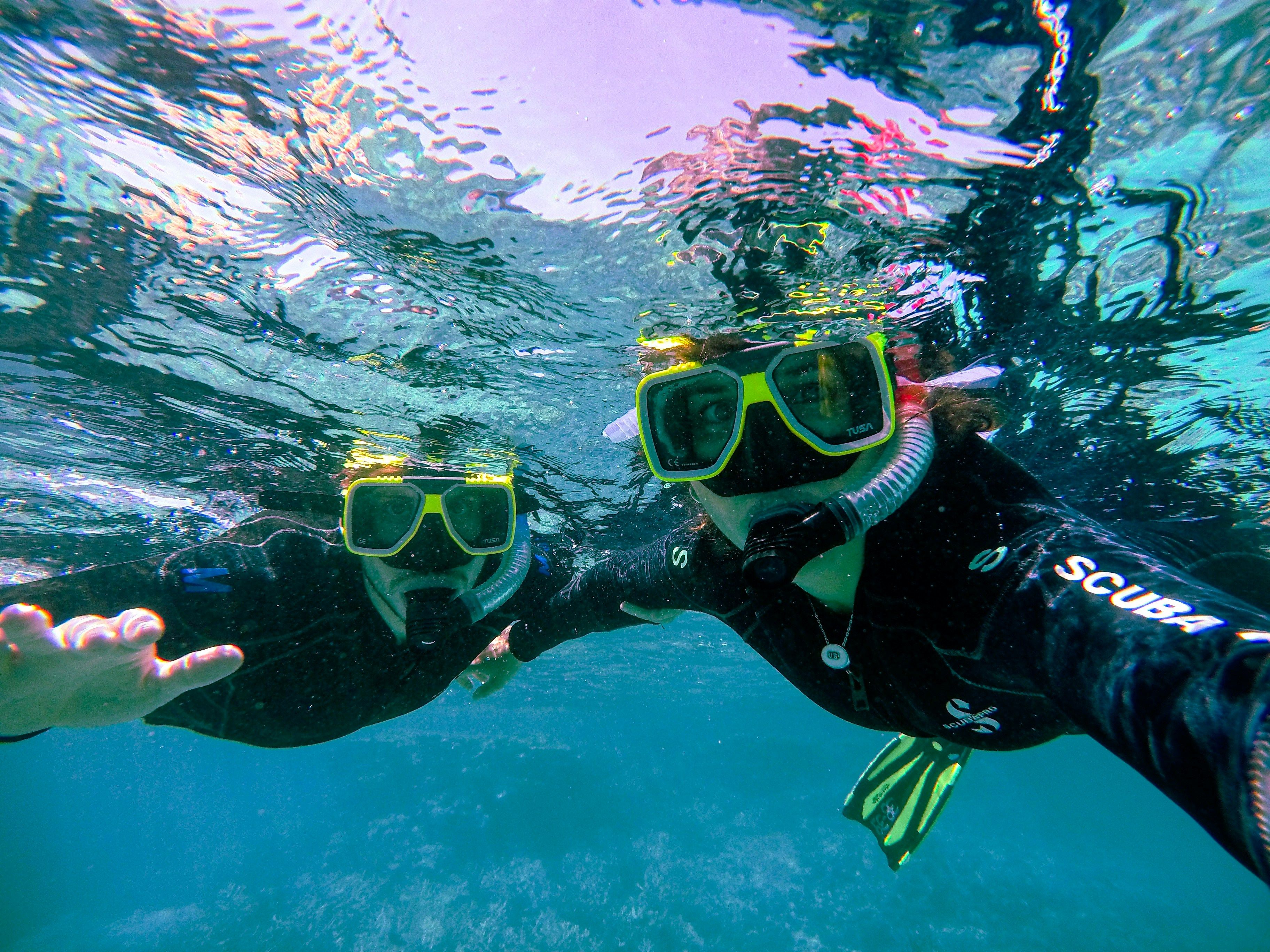 Two snorkelers underwater, wearing full gear and posing for a selfie while exploring the ocean