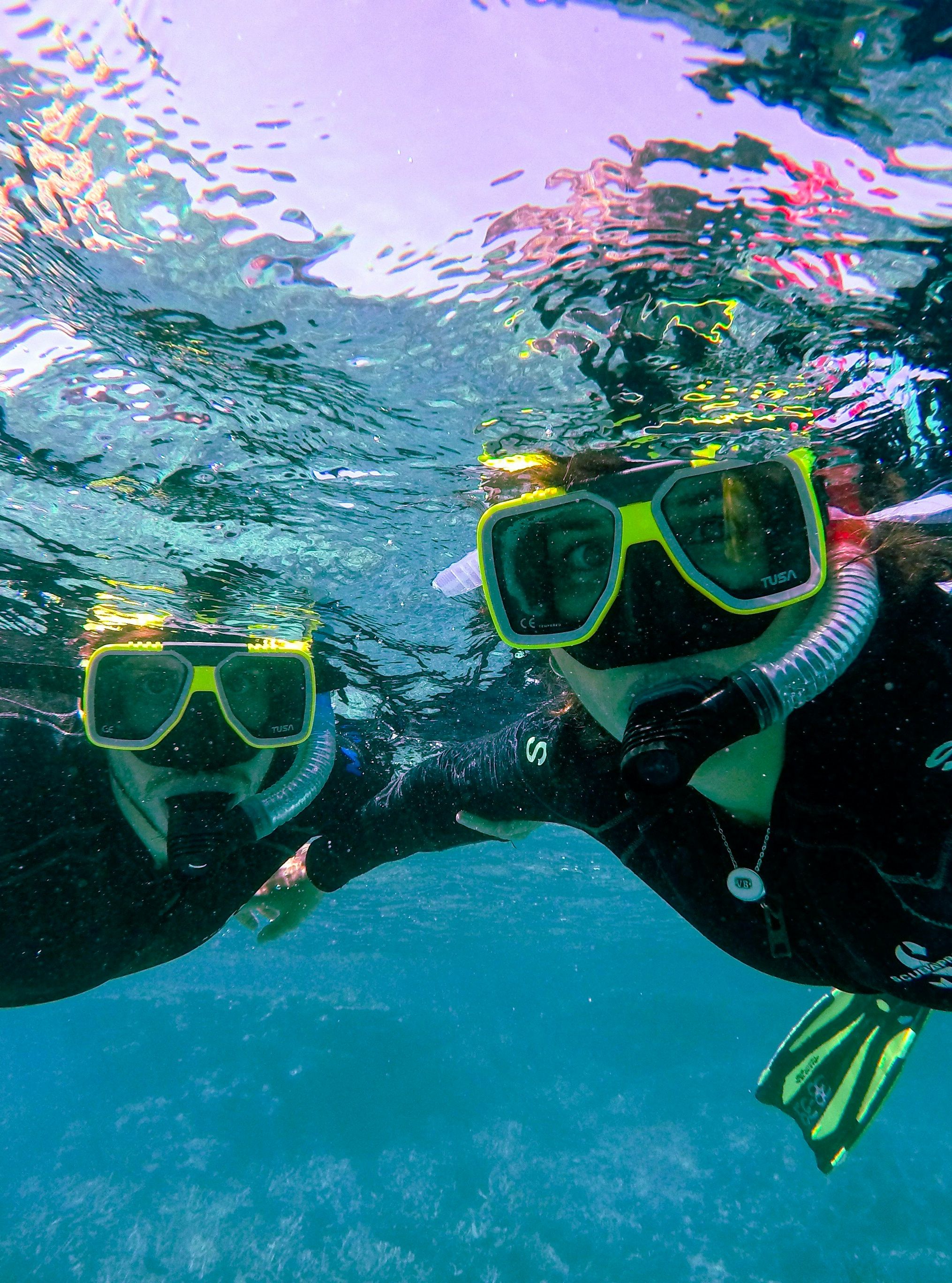 Two snorkelers underwater, wearing full gear and posing for a selfie while exploring the ocean