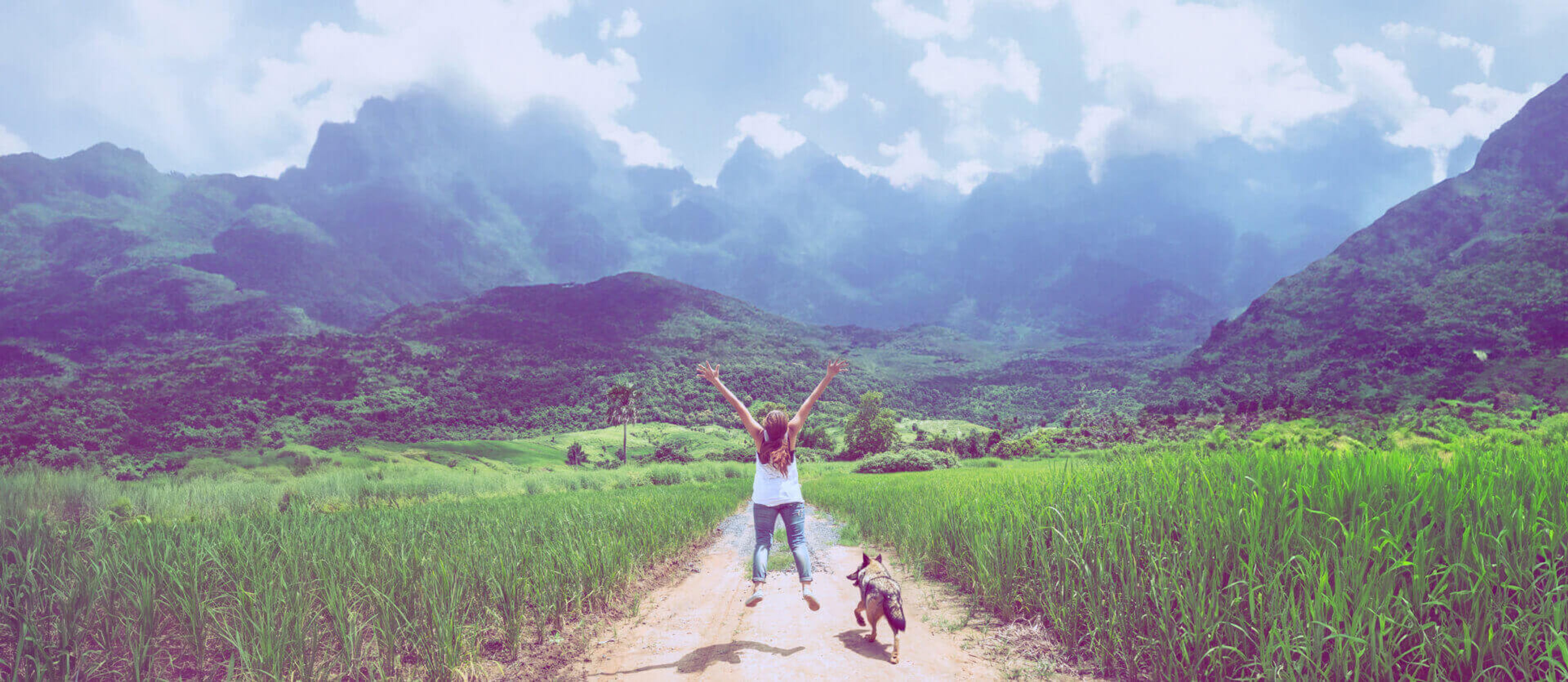 A joyful moment of a woman with arms raised, walking with her dog along a scenic pathway surrounded by lush greenery and dramatic mountain peaks under a partly cloudy sky.