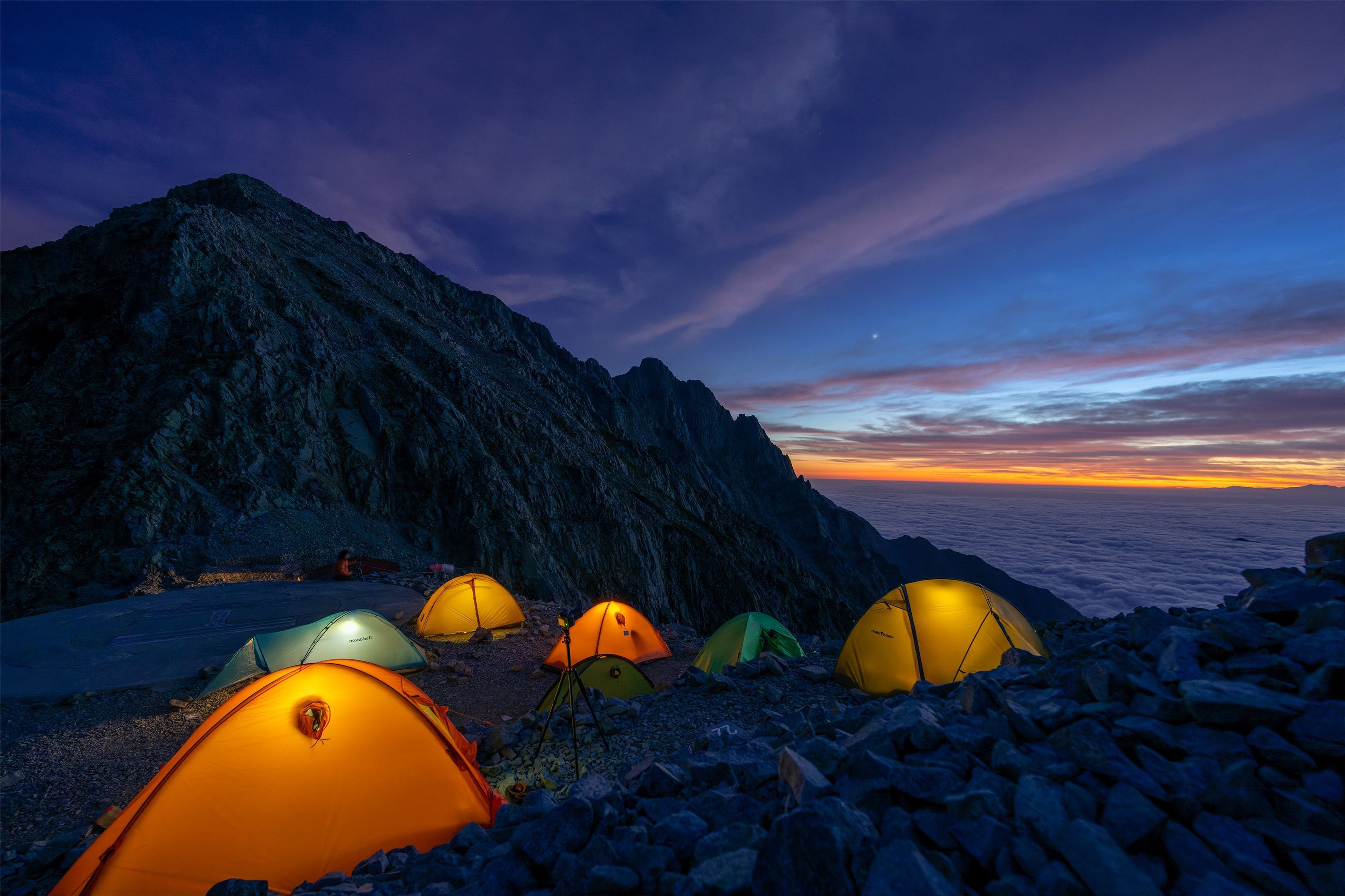Tents illuminated by lanterns at a campsite on a rocky mountainside during twilight, with a vibrant sunset in the background