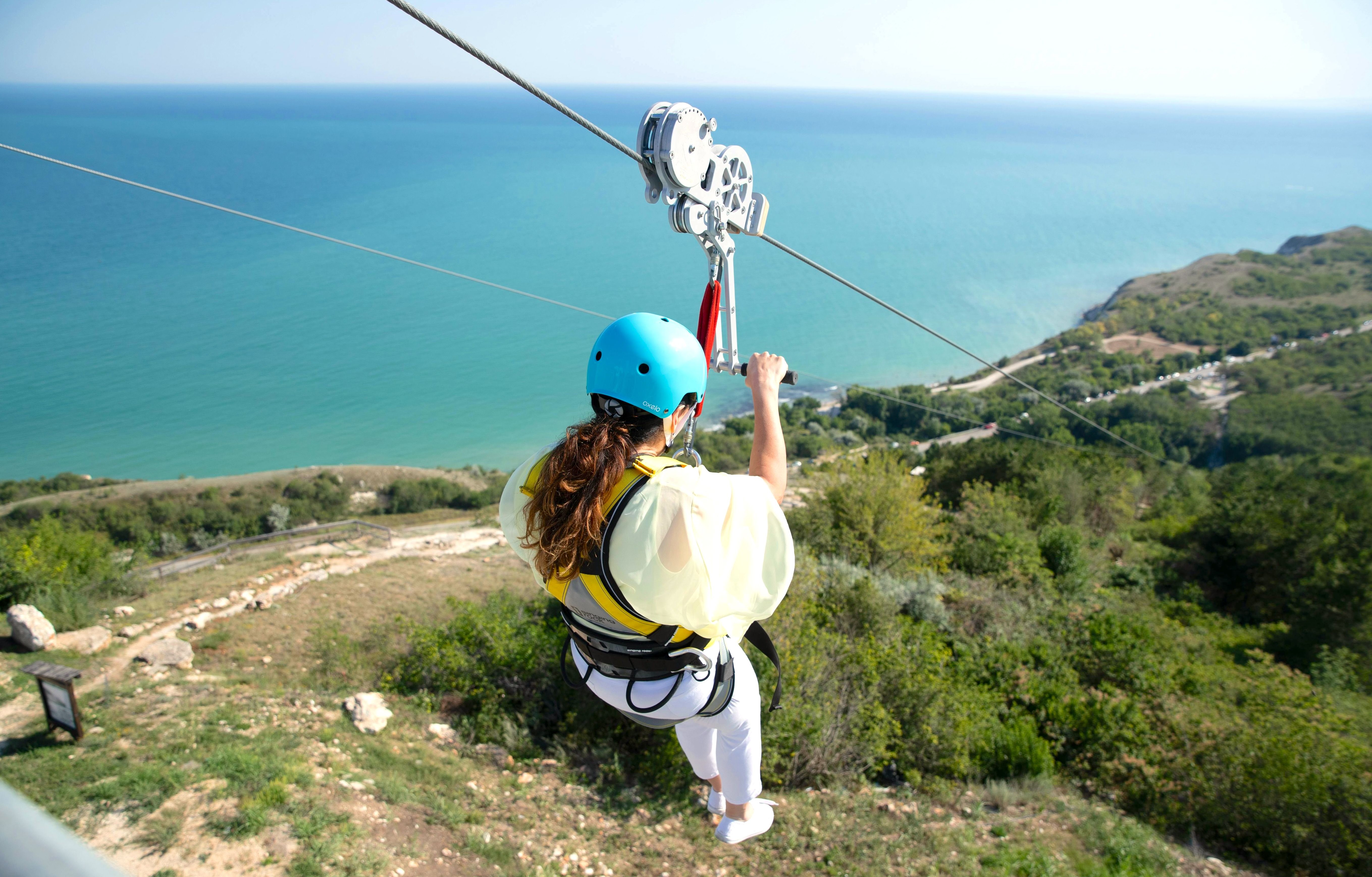 Person ziplining down a high hill with a scenic view of the sea and greenery in the background