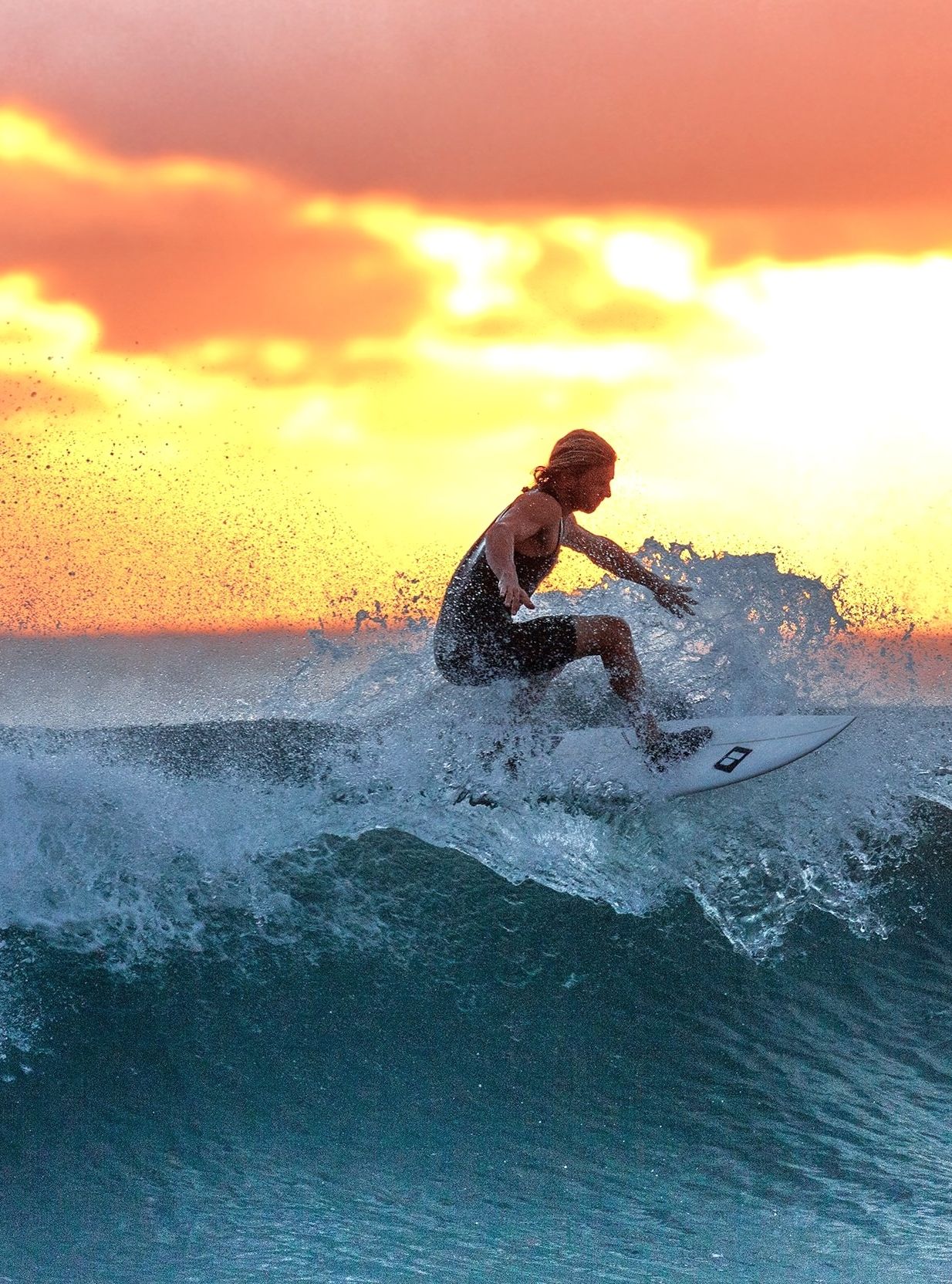 Surfer catching a wave during a vibrant orange sunset, with waves crashing beneath the board