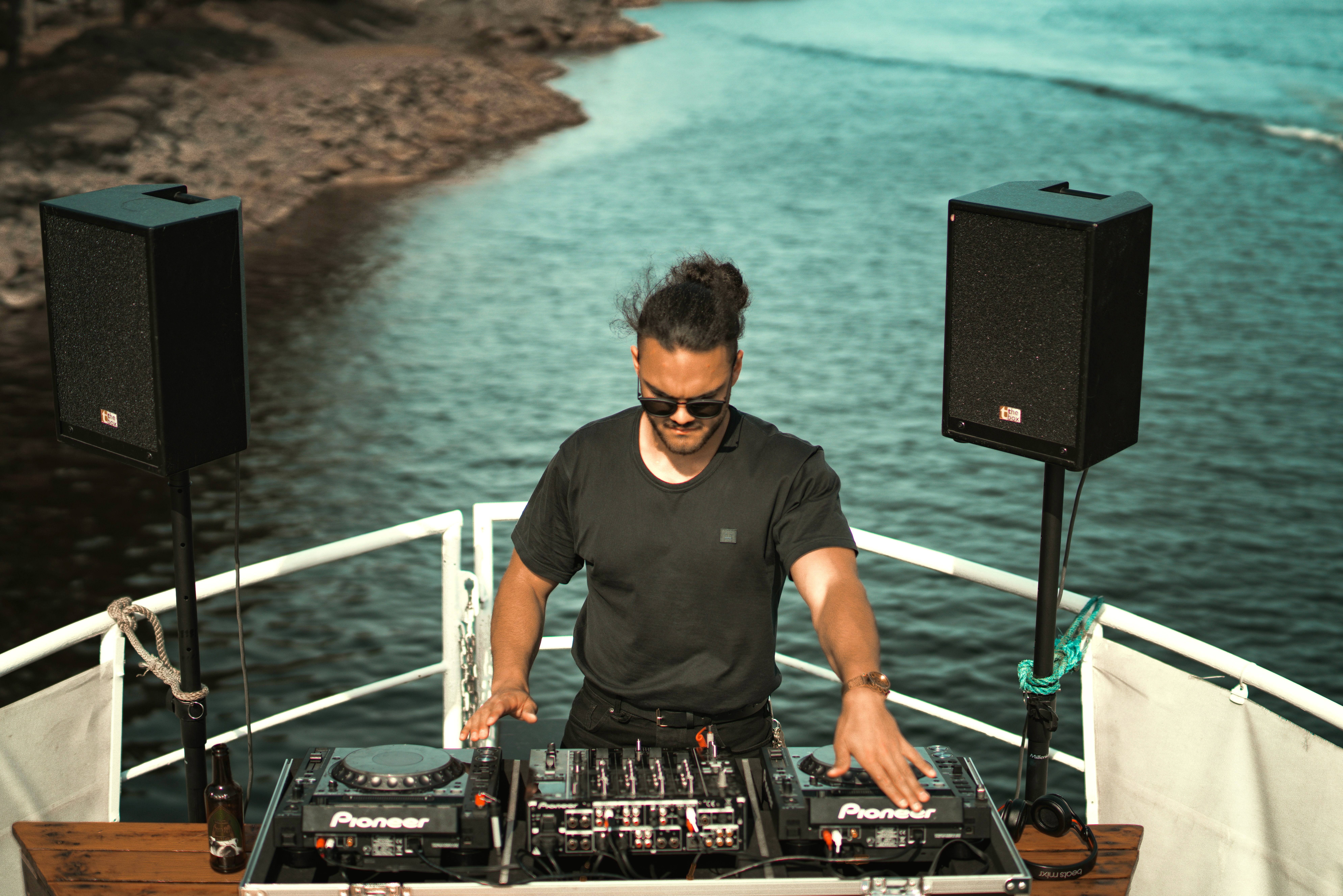 DJ performing on an outdoor deck with a sea in the background, surrounded by speakers and DJ equipment