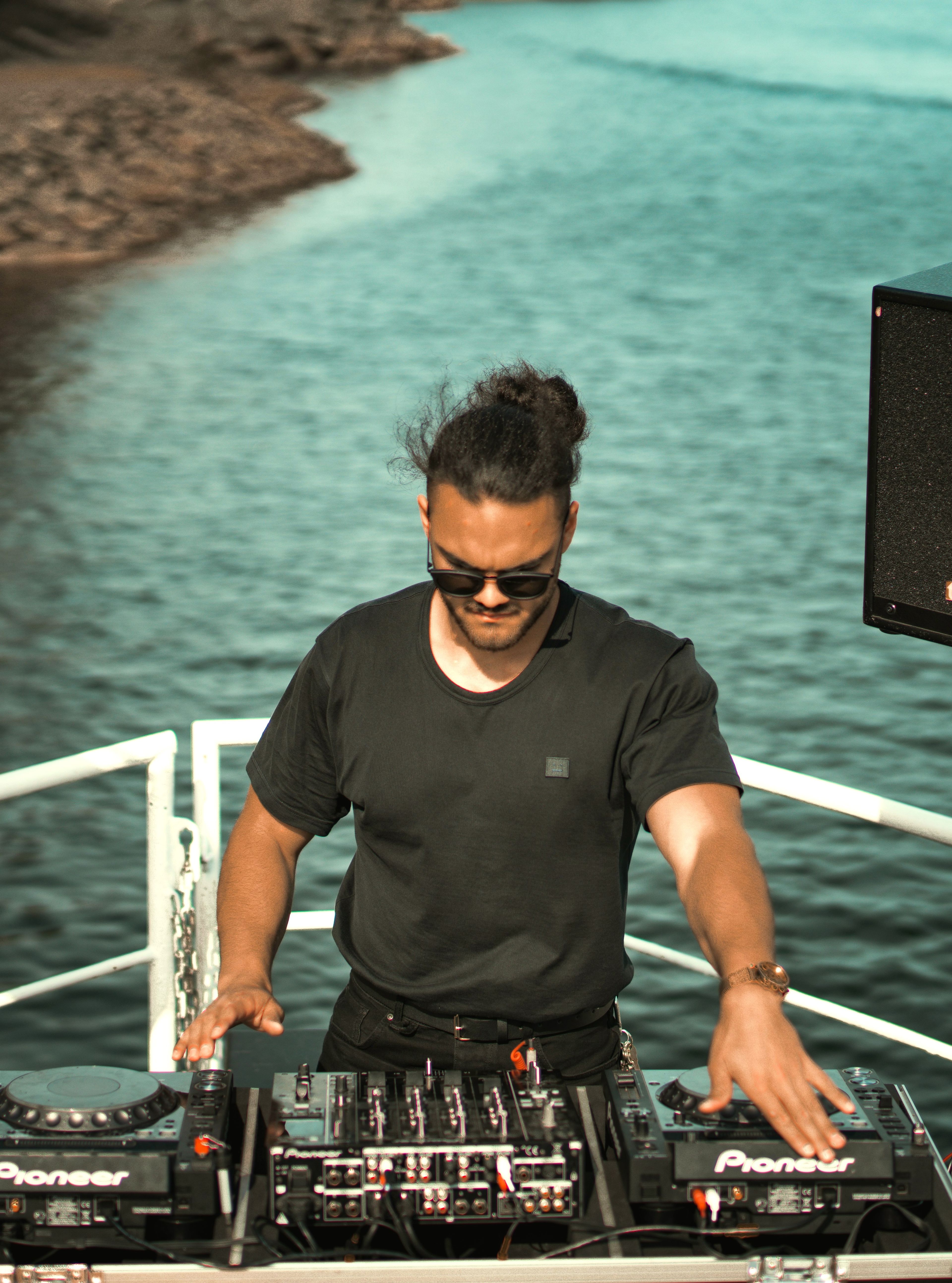 DJ performing on an outdoor deck with a sea in the background, surrounded by speakers and DJ equipment