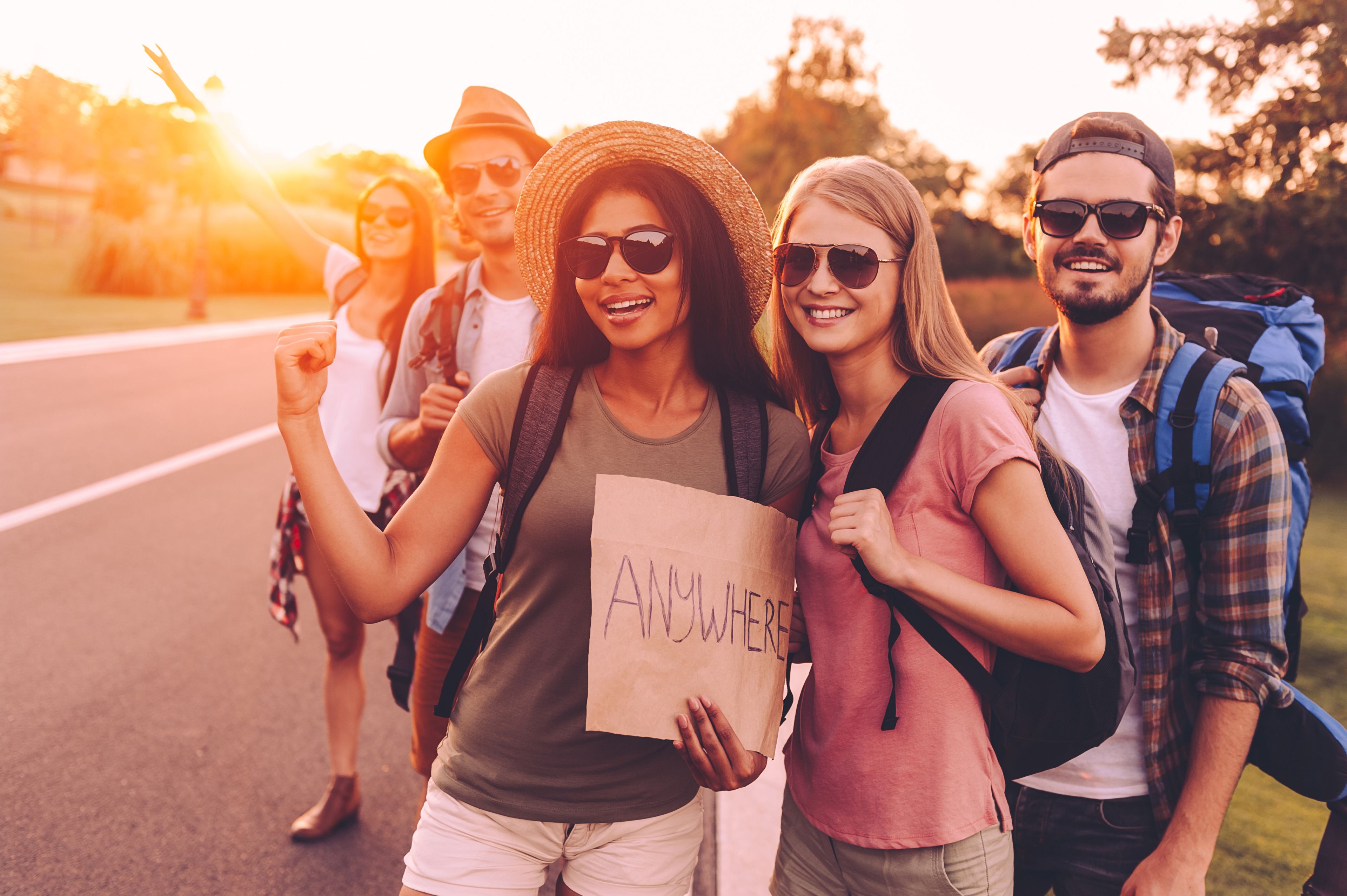 Group of smiling friends hitchhiking at sunset, holding a sign that says 'Anywhere'